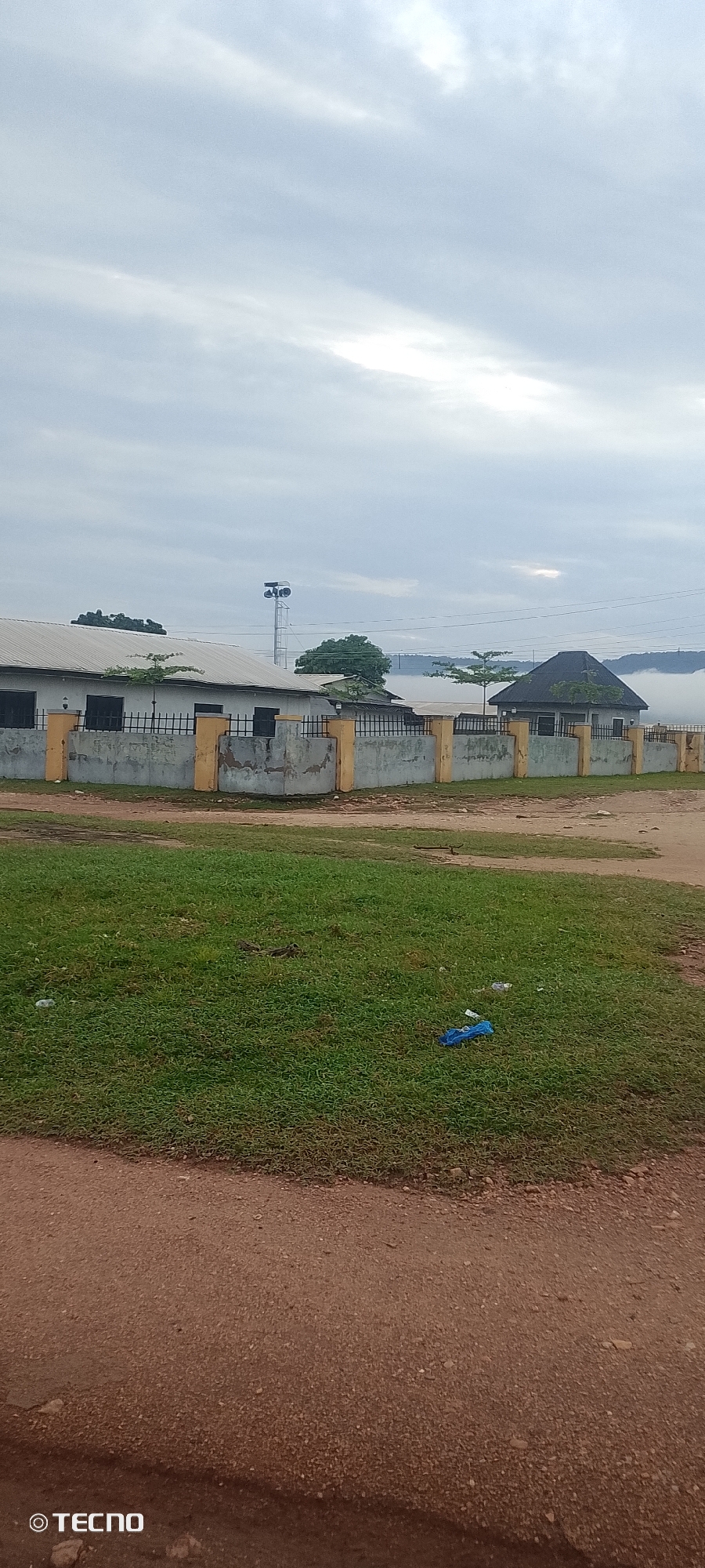 In the city of Adankolo, Nigeria, a low-angle shot captures a street scene under an overcast sky. A light grey concrete wall with yellow pillars runs horizontally across the mid-ground, marking the boundary of a property. Behind the wall, two single-story buildings with white roofs are visible. The building on the left has dark rectangular windows and a metal fence in front. To the right of this building, a tall metal tower with lights at its top stands prominently. Further to the right, another building with a dark, pitched roof is visible, partially obscured by mist or fog in the background, which also obscures a hilly landscape. In the foreground, a patch of vibrant green grass is present, with scattered litter including a blue plastic bag and some white plastic bottles. A dirt road runs along the bottom of the frame, leading into the foreground. The Tecno logo is present in the bottom left corner of the image.