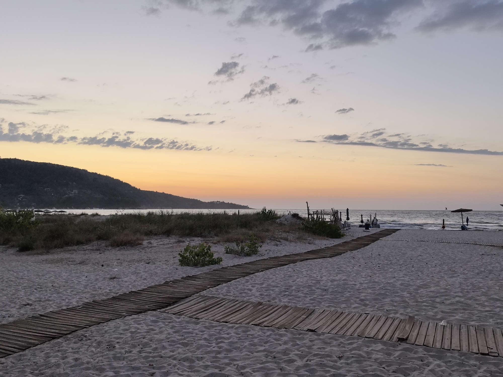 The image depicts a tranquil beach scene at dusk in Golden Beach, Greece. The sky transitions from a pale lavender at the top to a soft orange and yellow gradient near the horizon, with scattered clouds. A dark, tree-covered hill forms the background to the left. In the foreground, a sandy beach is crossed by two wooden boardwalks, one in the lower left and another extending towards the right and center. The boardwalks are constructed from parallel wooden planks laid on the sand. Beyond the boardwalks, sparse vegetation and beach umbrellas with chairs are visible, indicating a beach area. The sea is calm, with gentle waves lapping at the shore. The overall atmosphere is serene and peaceful.