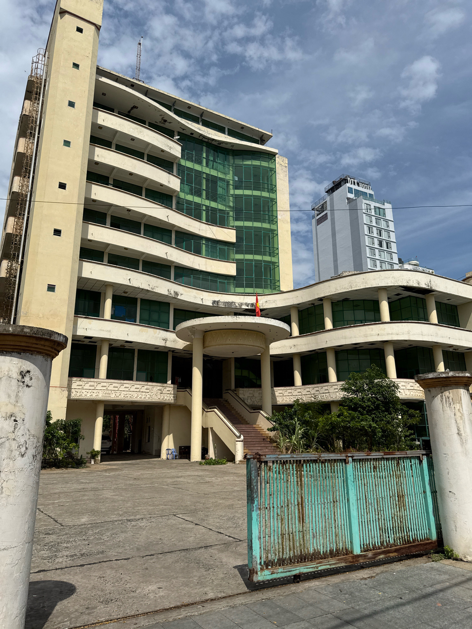 The image captures the exterior of a large, somewhat aged building complex in Nha Trang, Vietnam, under a cloudy sky. The primary building is a multi-story structure with a cream-colored facade, featuring curved balconies with green-tinted windows. A concrete ramp leads up to an entrance supported by columns. A weathered, turquoise metal gate is prominently positioned in the foreground. In the background, a modern, white high-rise building with the sign "DTX HOTEL" is visible. The area appears to be a paved courtyard with some sparse vegetation and trees. There are no people clearly visible in the scene. The overall impression is of a large, established property in a coastal city.