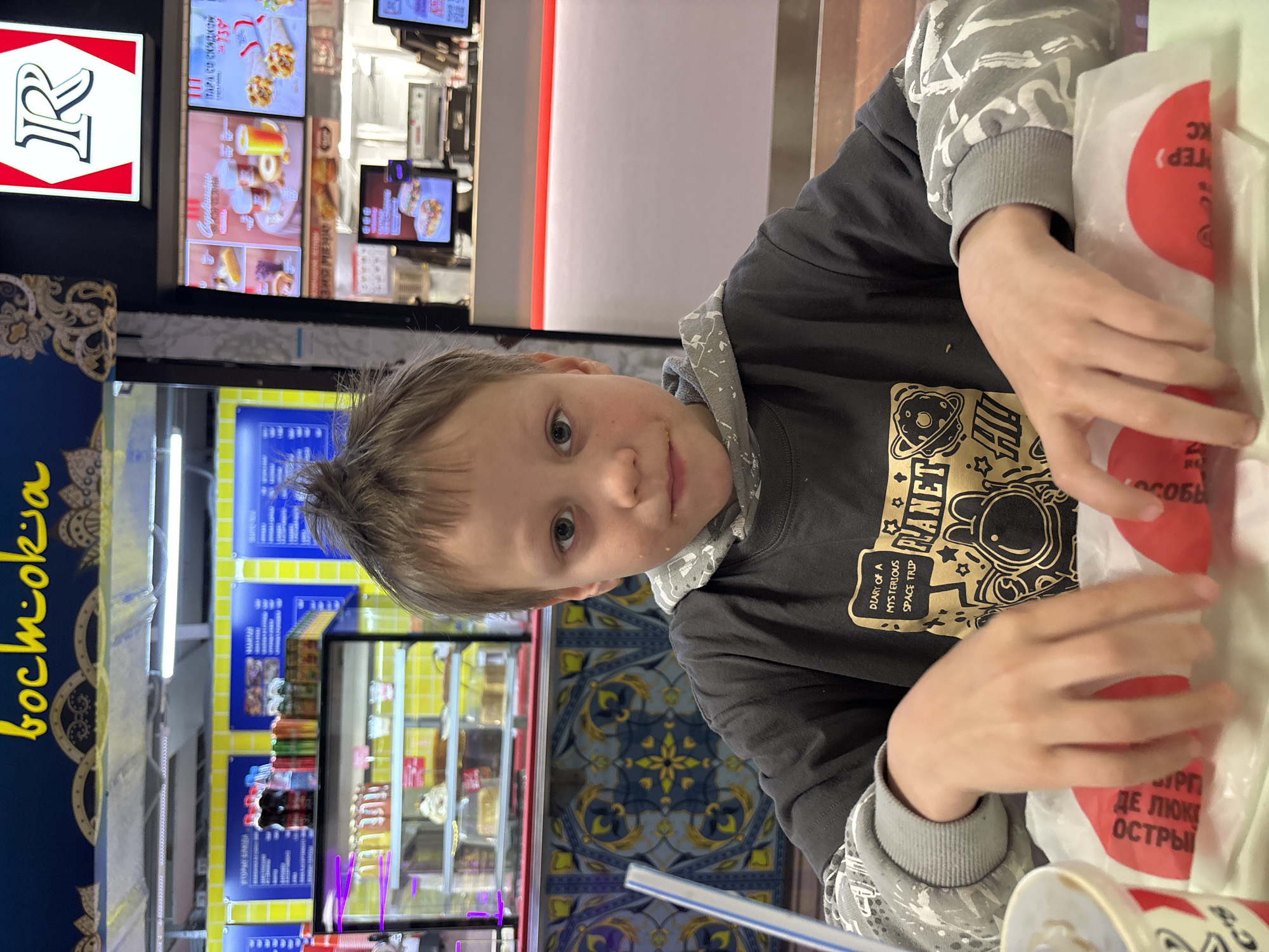 A young boy is sitting at a table in a fast food restaurant. He has light brown hair and blue eyes. He is wearing a dark gray hooded sweatshirt with a graphic print of an astronaut. He is holding a wrapped food item in his hands. The restaurant has blue and yellow decor. The restaurant name "bochmokia" is visible on the left side. In the background, a menu board with images of food and prices is visible, along with other customers and employees. The words "DIARY OF A MYSTERIOUS SPACE TRIP" and "PLANET" are visible on the boy's sweatshirt. The food item has red circles printed on its wrapping. The text "DE LOKE OSTRY" and "SOBBI" are visible on the wrapping. Based on the Cyrillic text and other signs, the location is likely in Grushovaya Balka, Russia.