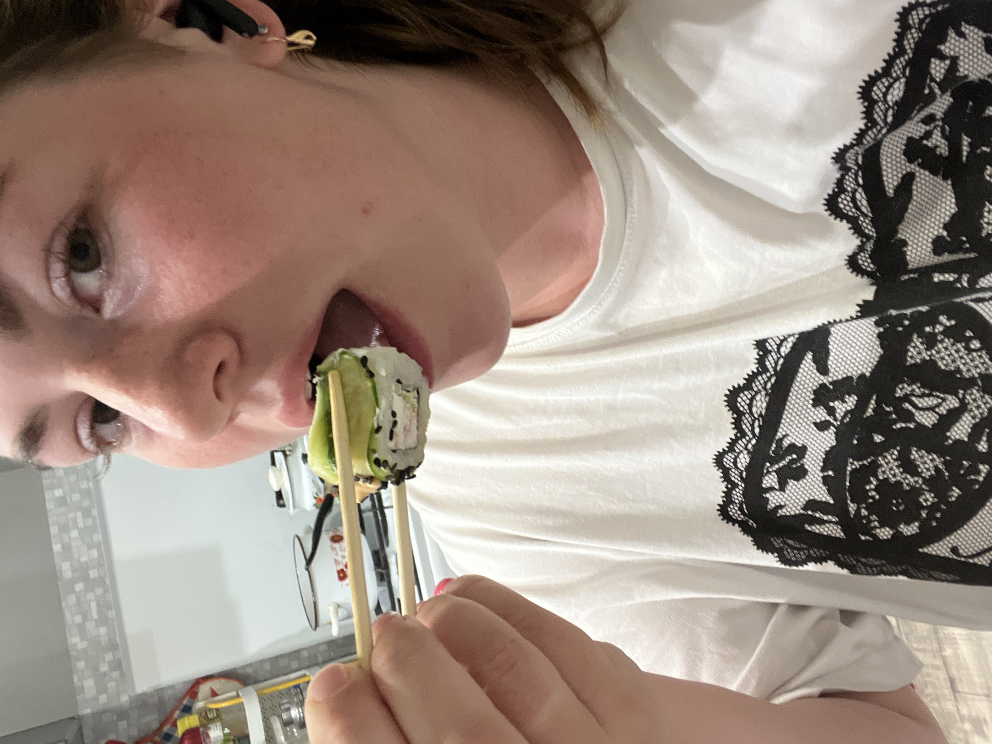 A fair-skinned person is holding a piece of sushi with chopsticks up to their open mouth. They are wearing a white t-shirt with a black lace design. The background appears to be a kitchen setting. The photo was taken in Chaikovsky, Russia.