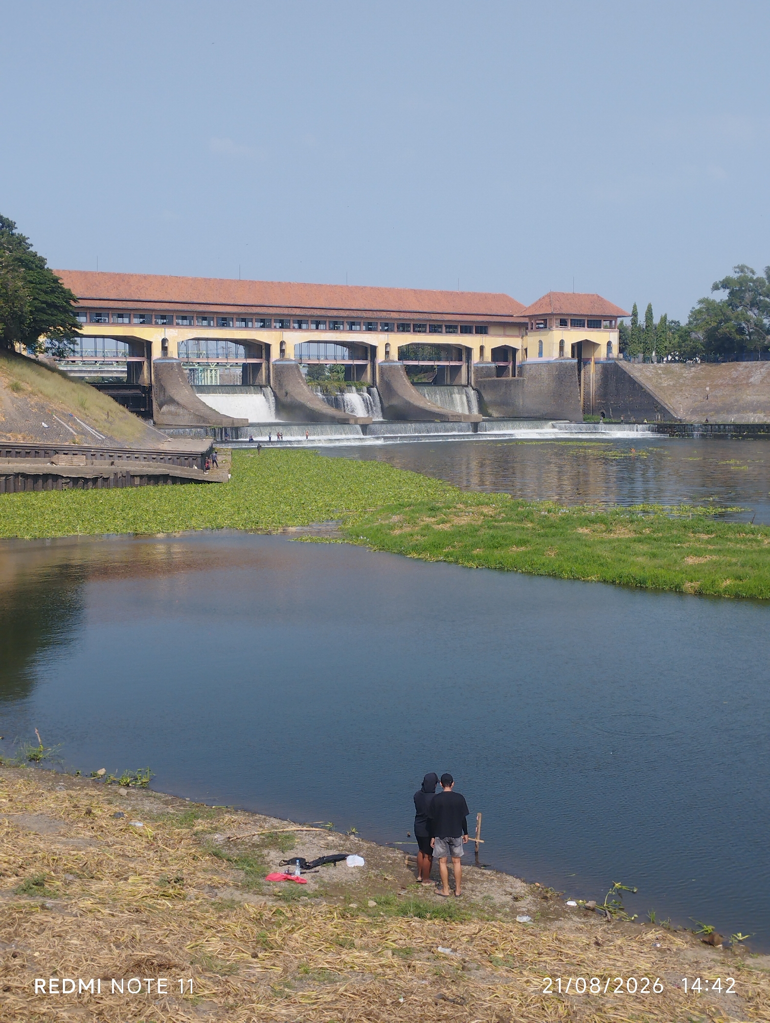 The image captures a scenic view of the Citarum River in Karawang, Indonesia, featuring a prominent barrage structure. The barrage, a long, yellowish building with a red-tiled roof and multiple arched windows, spans across the river. Water cascades over the barrage, creating a visible waterfall. The river itself is wide and reflects the clear blue sky. On the left bank of the river, a grassy slope leads up to the barrage, with trees on the far left. In the foreground, a patch of green aquatic plants floats on the water's surface. The right bank is mostly dry and covered with yellowish-brown vegetation. Two individuals, viewed from behind, stand on the dry bank near the water's edge, facing the river. One is wearing a black hooded jacket and black pants, while the other is in a black t-shirt and shorts. A few objects, including a red cloth, a black bag, and a white plastic bag, are scattered on the ground near them. The timestamp "21/08/2026 14:42" and "REDMI NOTE 11" are visible at the bottom of the image.