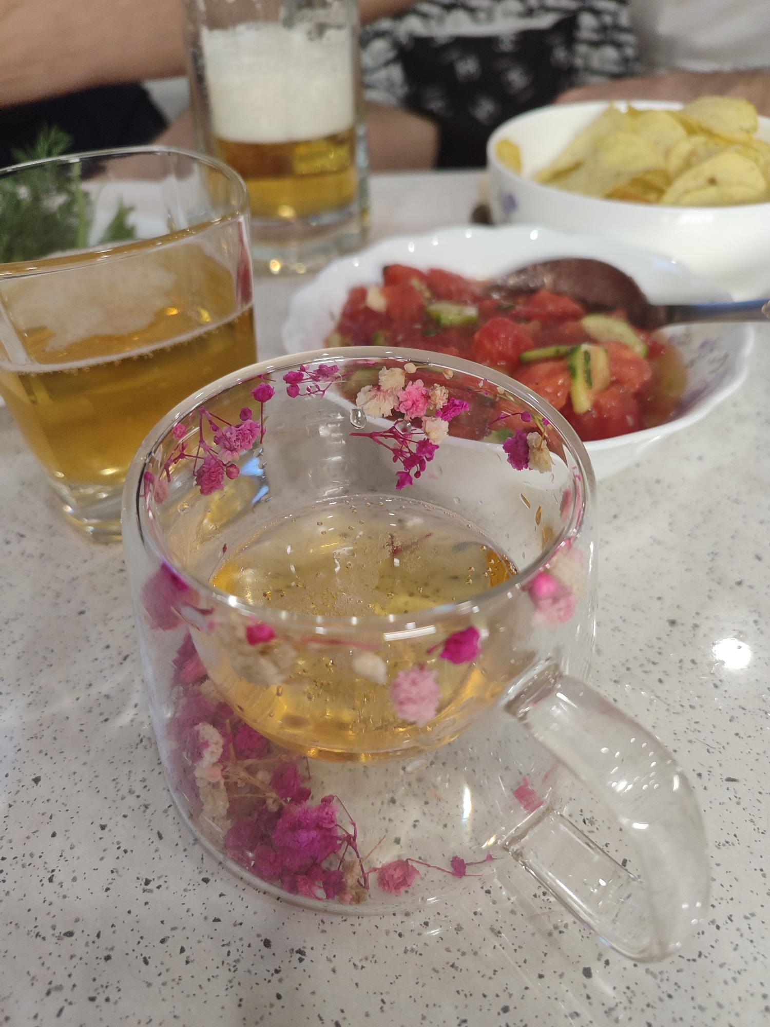 A close-up shot features a double-walled glass mug with delicate pink and white flowers embedded in its walls, filled with a pale yellow liquid, likely tea or a light-colored beverage. The mug is set on a speckled gray countertop. In the background, a tall glass of beer with a foamy head is visible, alongside a white bowl filled with potato chips. Another white bowl, in the middle ground, contains a salad of chopped tomatoes and cucumbers, with a spoon resting in it. The setting appears to be a casual meal or gathering, possibly indoors given the lighting. The overall mood is relaxed and convivial. The image has a shallow depth of field, with the flower-decorated mug in sharp focus and the background elements softly blurred. No visible text is present.