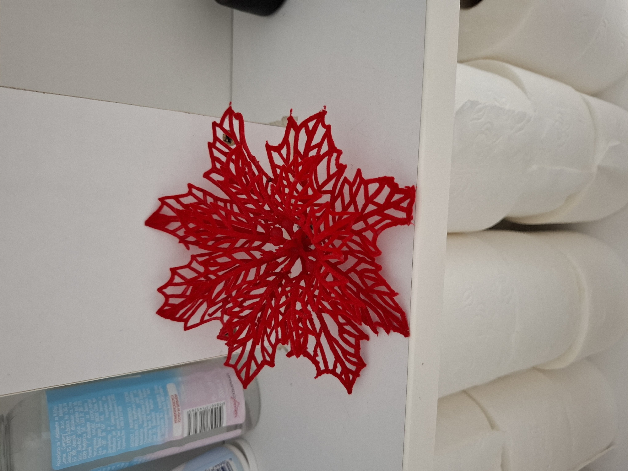 An overhead, close-up shot captures an indoor scene dominated by a red, filigree-style decoration resembling a poinsettia or snowflake. The decoration is positioned on a white shelf or surface, serving as a bright focal point against the neutral background. To the right, a stack of white toilet paper rolls is neatly organized on an adjacent shelf, partially visible. At the bottom left, a clear bottle with a blue label is partially visible, likely containing hand soap or lotion. The overall setting suggests a domestic storage area, possibly a bathroom or utility closet, characterized by its clean white surfaces. The lighting is even, typical of indoor illumination.
Visible text from the bottle label includes distribution information in various languages, mentioning "Proizvedeno za i distributer za", "Lomax Company", "Subotica", "Podgorica", and "Hercegovine MALISIC EXPORT". A prominent phrase, "DERMATOLOGICALLY TESTED IN GERMANY," is also visible, along with a barcode starting with "8606012716383". No activity is taking place, and there are no people present.