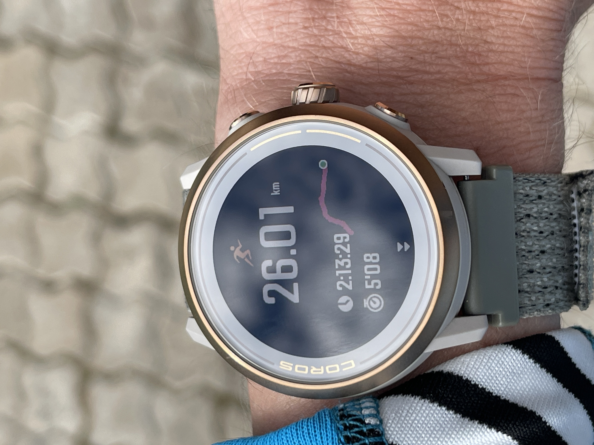 A close-up shot shows a wrist wearing a Coros brand smartwatch. The watch face displays running metrics: 26.01 km distance, 2 hours 13 minutes and 29 seconds of activity, and a pace of 5 minutes and 8 seconds per kilometer. The watch band is gray with a woven texture. A striped blue, white, and black fabric is visible near the bottom right of the frame, possibly part of clothing. The person's skin tone is fair. The location is not determinable from the image.