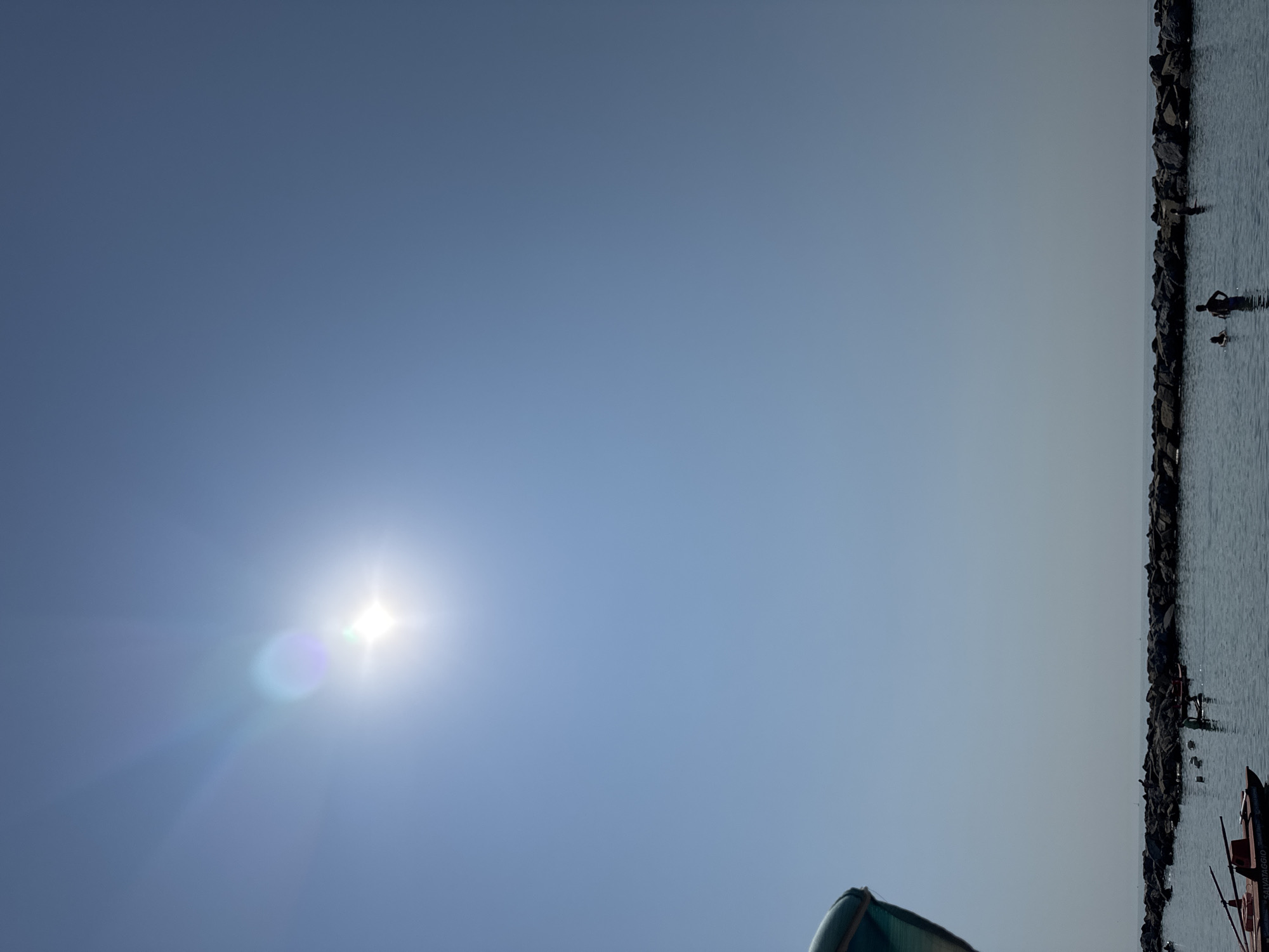 The image depicts a bright, sunny day at a beach in Sanremo, Italy, likely near the port area given the presence of a rocky breakwater and a small boat. The sun is high in the clear blue sky, creating lens flare and rays. In the water, several people are visible. Some are wading or swimming near the breakwater, while others appear to be on paddleboards or in small boats. The breakwater itself is made of large, dark rocks, and it extends horizontally across the mid-section of the image, separating the calmer water in the foreground from the open sea beyond. A portion of a green awning or umbrella is visible in the lower left corner, suggesting a beachside setup. The water is calm and reflects the bright sky. The overall impression is one of a typical summer day at the beach.