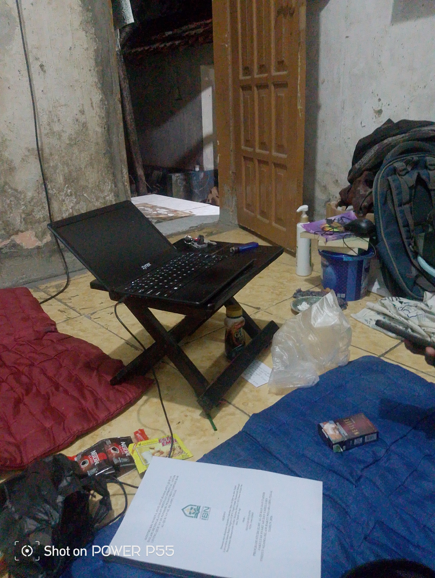 The image depicts a cluttered indoor scene, likely a simple dwelling or temporary living space. A laptop is set up on a portable folding stand in the center, positioned on a tiled floor. To the left of the laptop, a dark red sleeping mat is visible, and to the right, a blue tarp covers part of the floor. Various items are scattered around, including what appears to be a backpack, plastic bags, bottles, and packets of instant coffee. A pack of cigarettes rests on the blue tarp in the foreground. The walls are bare and appear to be made of concrete, with some signs of wear and tear. A wooden door is visible in the background. The overall impression is one of a utilitarian and somewhat disorganized living area. The location is identified as Lebaksiu, Indonesia.