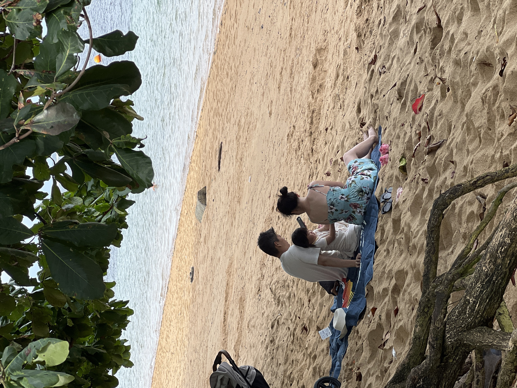 The image shows a family of three—a woman, a man, and a small child—lying on a blue blanket on a sandy beach in Denpasar, Indonesia. The woman is wearing a floral swimsuit, and her hair is in a bun. The man is wearing a white t-shirt, and the child is wearing a light-colored top. They appear to be looking at a phone or device that the man is holding. A baby stroller is partially visible in the bottom left corner of the image. There are trees and foliage framing the scene on either side. The ocean is visible in the background along the shoreline.