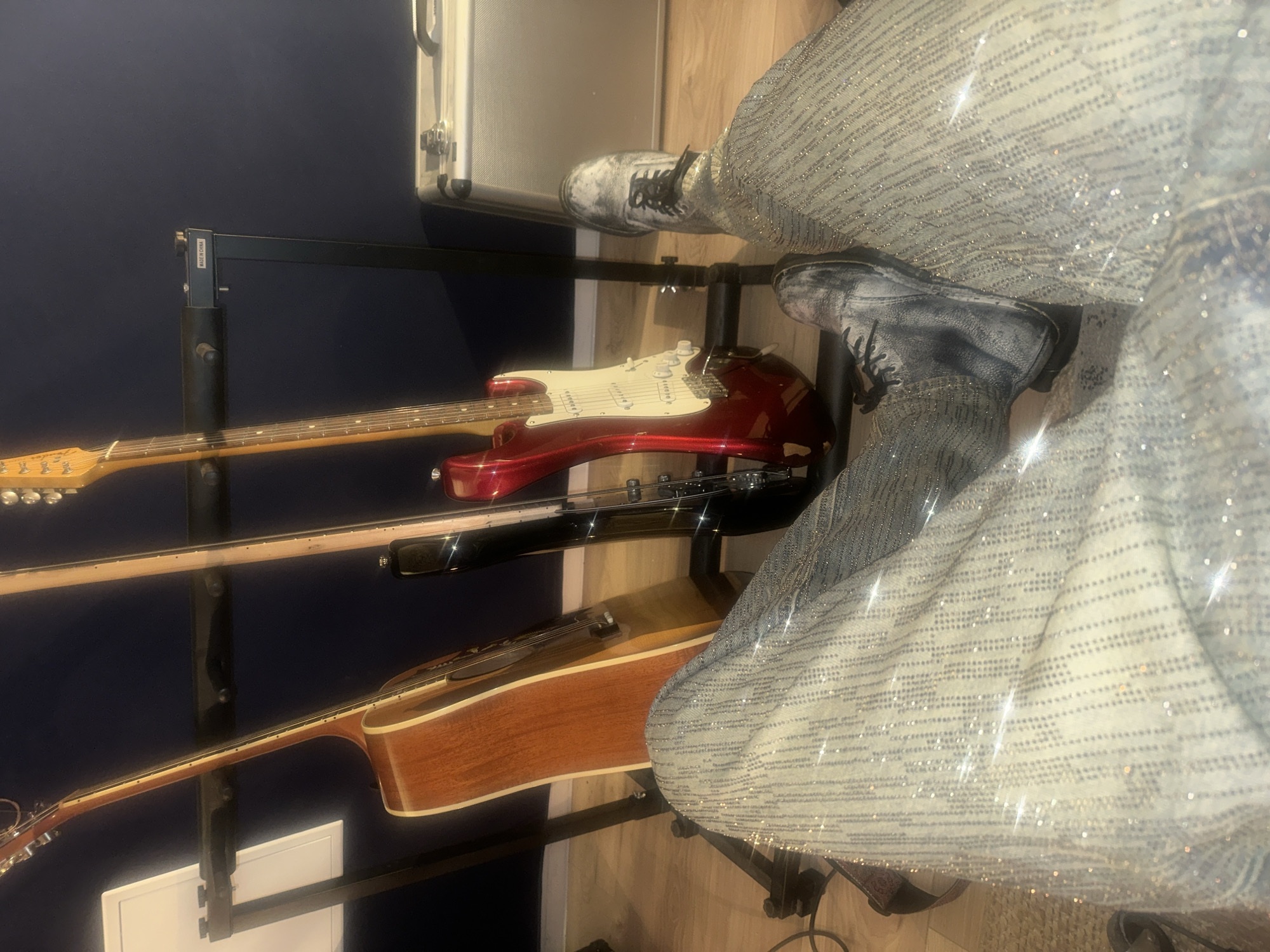 The scene depicts an indoor setting within the city of Paris, France, resembling a music studio or practice room. The perspective is a low-angle, first-person view, showing the lower extremities of one individual. The person's legs are clad in light-colored trousers heavily embellished with small, reflective studs or rhinestones arranged in vertical rows. Their feet are covered by distressed grey and black lace-up boots with black soles. In the background, three stringed instruments are displayed on a black multi-guitar stand: a natural wood acoustic guitar, a black electric bass guitar, and a red electric guitar with a white pickguard. A label reading "MADE IN CHINA" is visible on the guitar stand. To the right of the instruments, a silver metallic equipment case or amplifier rests on the floor. On the far left, a black music stand holds a white sheet or board. The floor is constructed of light-toned wood, and the wall behind the instruments is dark blue. The individual is stationary, with no active interaction with the instruments depicted.