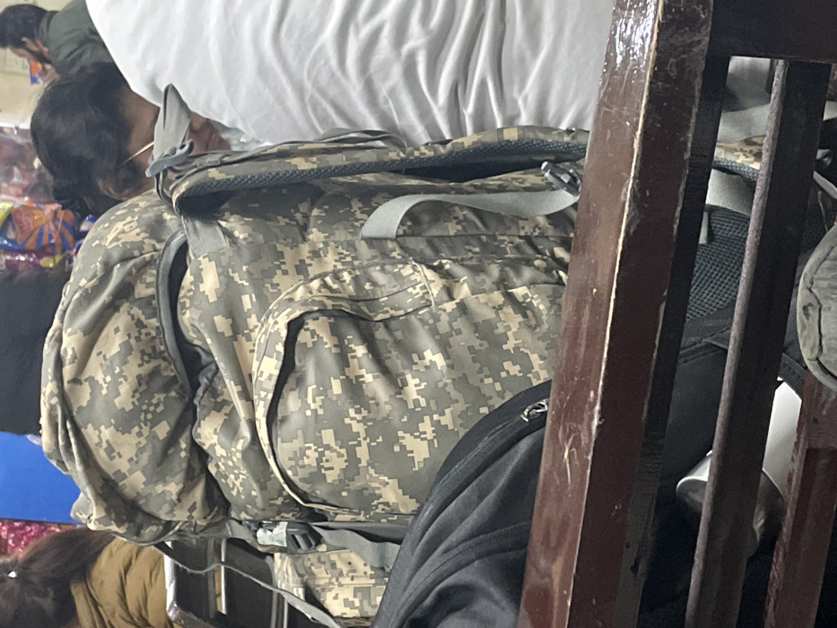 The image captures a partial view of a person wearing a large, digital camouflage backpack, characteristic of military or tactical gear, possibly a Universal Camouflage Pattern (UCP). The individual is seen from behind, appearing to be seated or resting in an indoor setting. Their head is partially visible with dark hair and what may be glasses or an earpiece.
Above the backpack is a white, soft item, likely a pillow or blanket, suggesting a resting or sleeping arrangement. To the right, a dark wooden structure, possibly part of a bed frame, bunk bed, or shelving unit, is prominent. Another dark-colored bag is partially visible to the right of the camouflage pack. In the blurred background, there are colorful, indistinct objects that could be packaged goods or other belongings, and a second person with dark hair is faintly discernible.
The scene appears to be dimly lit, indicative of an indoor environment with low illumination, possibly in a crowded space such as a dormitory, train, bus, or temporary shelter. No specific active event is occurring; the image depicts a moment of rest or transit. There is no visible text in the image. The location context for this scene is Haldwani, India.