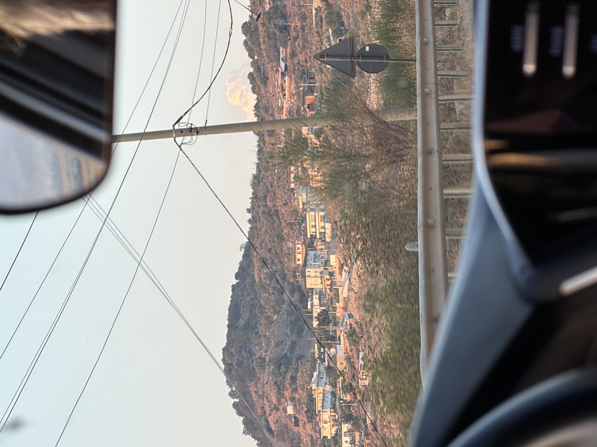 The image shows a hilly landscape with a cluster of buildings, likely a village, situated on the slopes. The buildings are primarily made of light-colored materials and appear to be residential. Vegetation, characterized by dry grasses and scattered trees, covers the hills. Several utility poles with wires crisscross the scene, suggesting a populated area. A traffic sign, a black triangle pointing upwards with a circular black sign below it, is visible on the right side of the image. In the foreground, the interior of a car is visible, including part of the dashboard and a side mirror reflecting a person. The sky is clear with a pale blue hue, and a fluffy white cloud is present in the background, above the village. The overall lighting suggests it is either early morning or late afternoon.