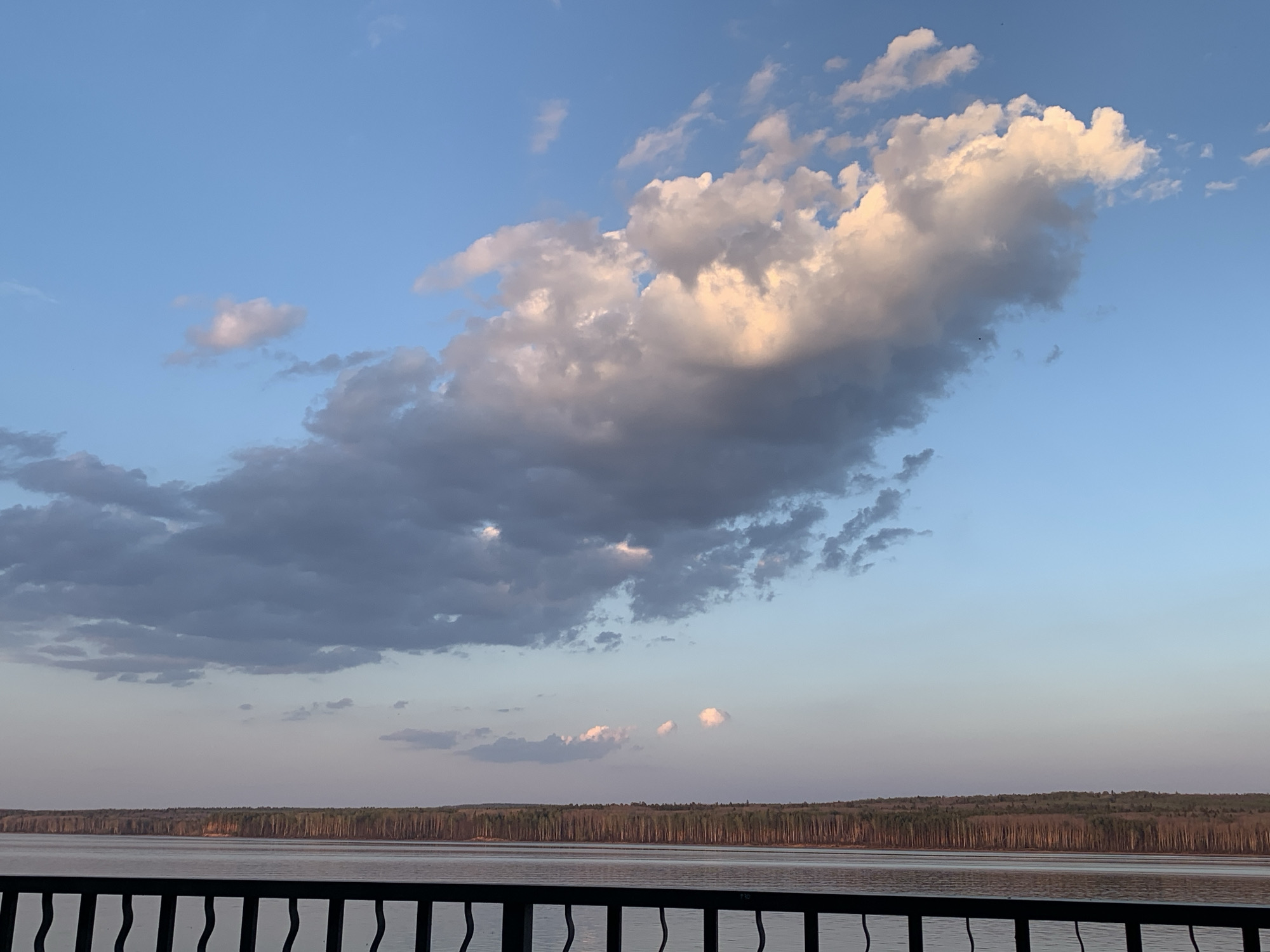 The image shows a view of the Kama Reservoir from Chaikovsky, Russia. The foreground features a dark metal railing. Beyond the railing is the reservoir with a calm surface. A forested shoreline stretches across the middle ground. Above, the sky is blue with varying shades, featuring cumulus clouds, some of which are white and illuminated, and others that are gray and shadowed.