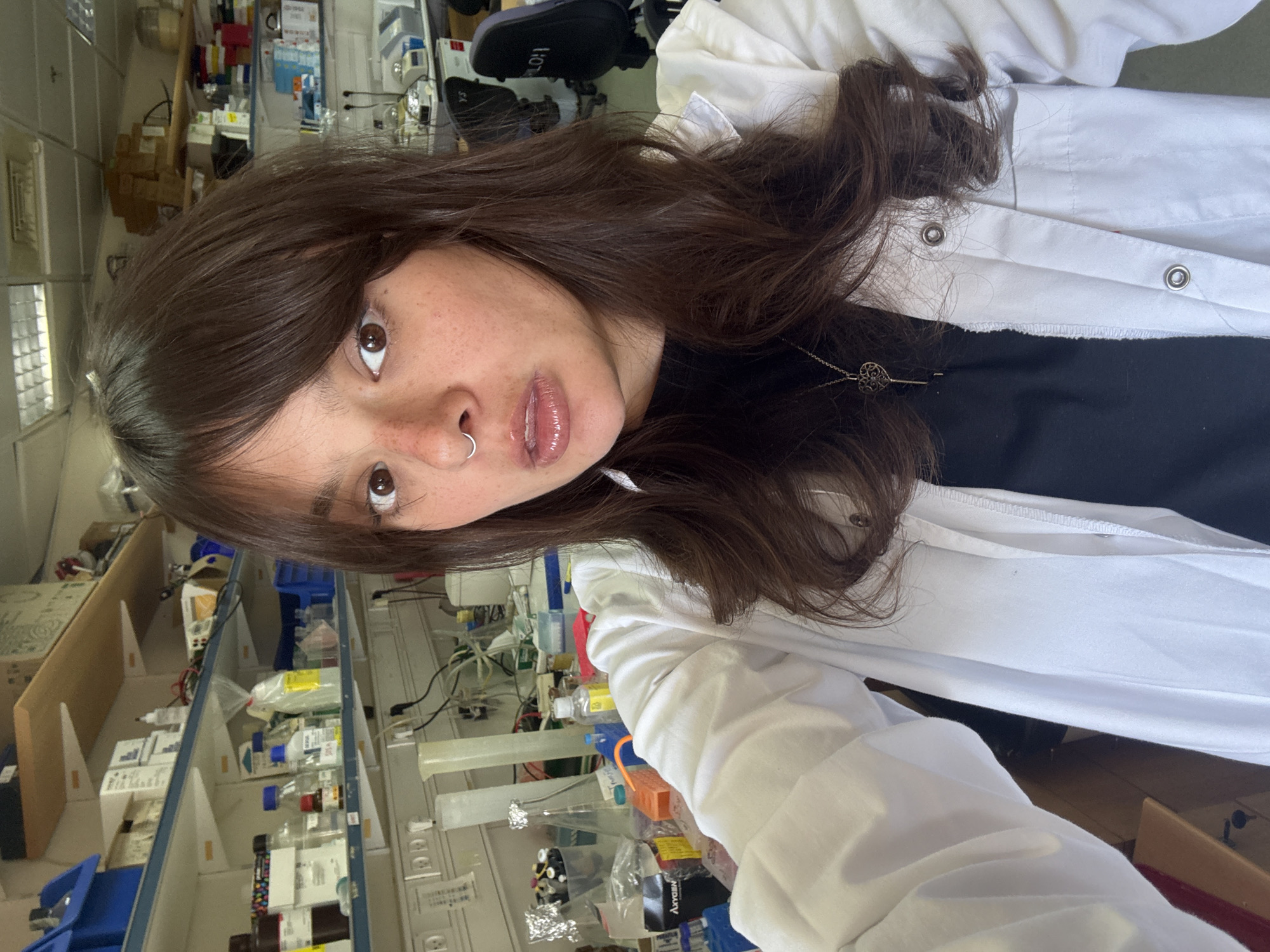 A young person with long, dark brown hair, wearing a white lab coat over a black shirt, poses for a selfie in what appears to be a laboratory. The person has light brown eyes, freckles across their nose and cheeks, and a silver hoop nose ring. Their lips are slightly parted, appearing to be wearing lip gloss. A delicate silver necklace with a key-shaped pendant is visible.
The background of the image shows rows of shelves filled with various laboratory equipment and supplies, including bottles, containers, and what look like scientific instruments. The lab appears well-lit, suggesting it could be daytime. The overall impression is that of a busy research or academic environment. The image is taken from a slightly high angle, looking down at the subject. No specific weather conditions or time of day can be definitively determined beyond the artificial lighting of the lab. The location context provided is Tel-Aviv, Israel. Some text is visible on items in the background, but it is not legible enough to be transcribed.