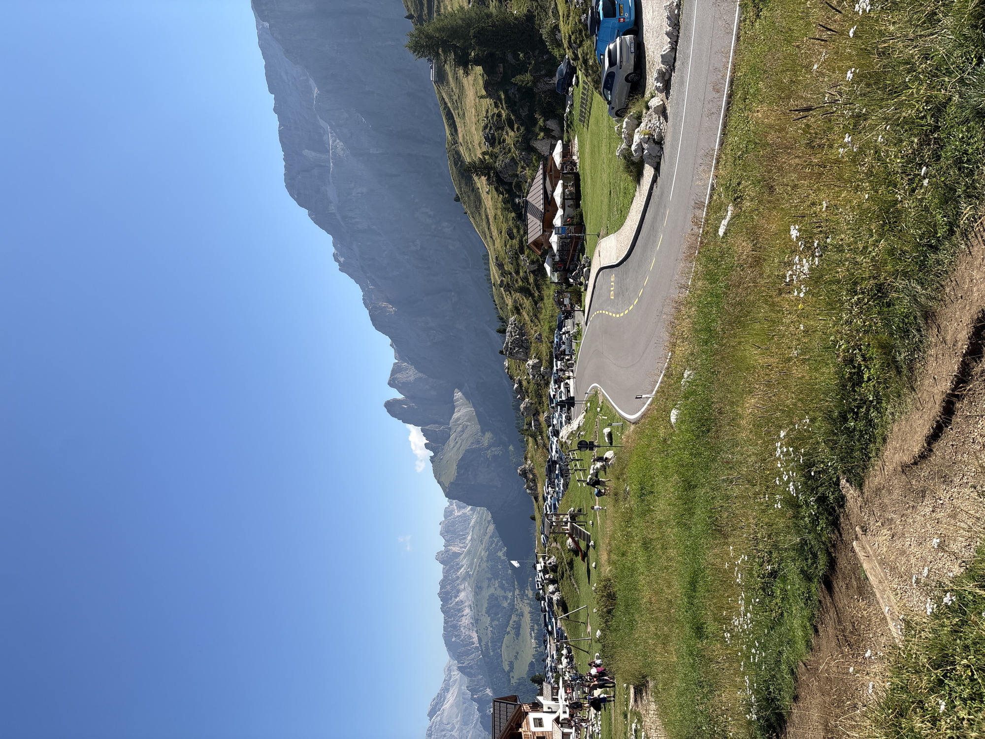 A panoramic, eye-level, outdoor, daytime shot captures a mountain landscape with a road and buildings in the foreground. The sky is a clear, bright blue. In the mid-ground, a paved road curves to the right, with a yellow line painted down its center and a white line along its edge. A sign with "BUS" is visible on the roadside. A large parking lot filled with numerous cars is situated to the left of the road. A playground structure with swings and a slide is also present in this area.
On the left side of the frame, a rustic building with a dark roof and wooden siding is partially visible. Several people are seen walking around this building and in the parking area. To the right of the road, another building with a similar rustic architectural style, featuring a dark roof and white umbrellas set up outside, appears to be a restaurant or cafe. Several parked cars, including a silver SUV and a blue van, are located near this building.
The landscape is characterized by rolling green hills covered in tall grass and scattered wildflowers. In the background, majestic, rugged mountains with steep slopes and jagged peaks dominate the horizon. The distant mountains are hazy, suggesting a significant distance. The overall scene depicts a busy tourist area in a mountainous region on a sunny day.
