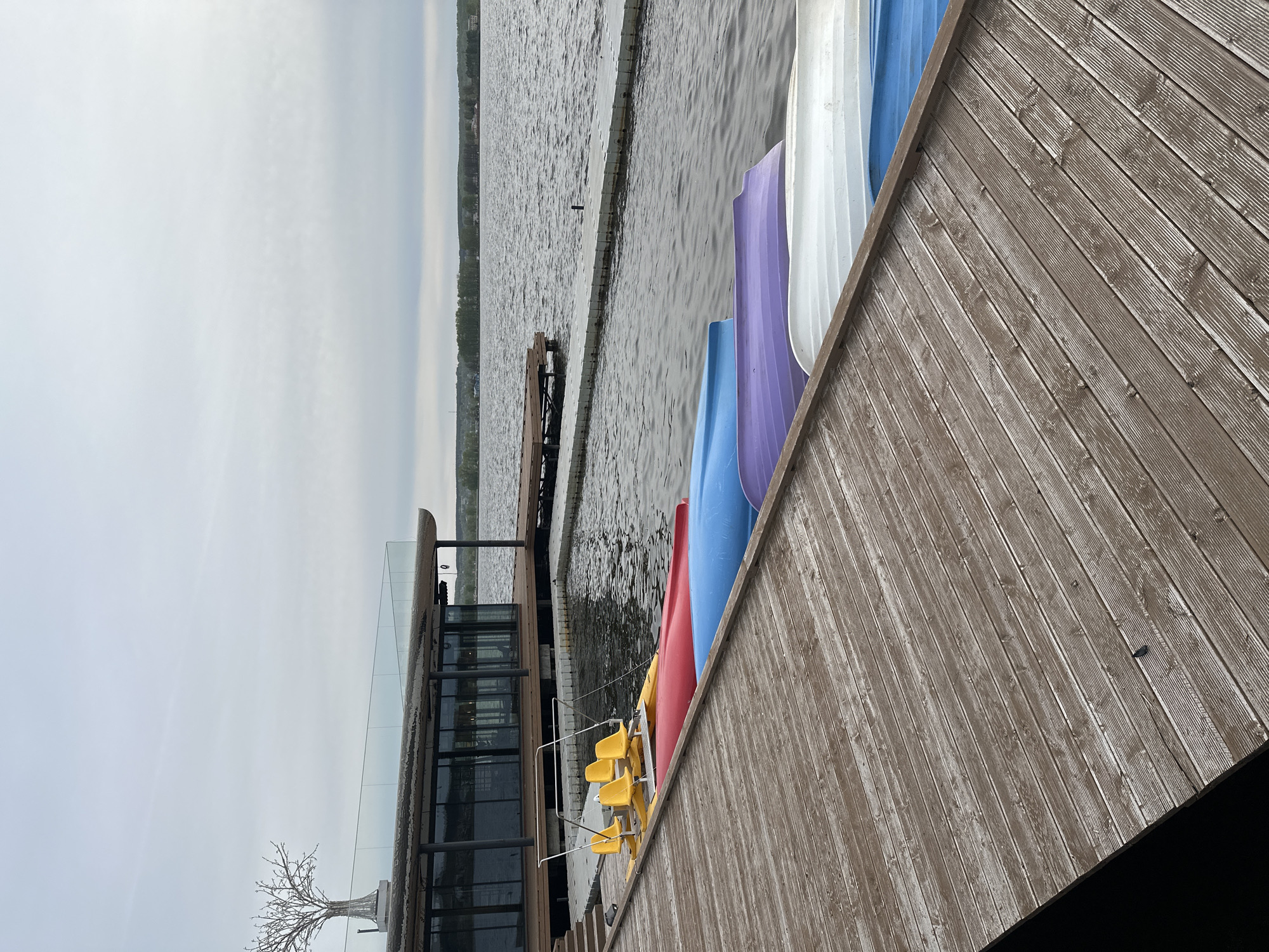 The image depicts a scene on a wooden pier in Votkinsk, Russia. Several colorful kayaks (red, blue, purple, white) are stored on the right side of the pier. Yellow chairs and a modern building with glass panels are visible on the left. In the background, a body of water stretches towards the horizon, with a distant shoreline visible. A small, partially submerged dock extends into the water. The sky is overcast.