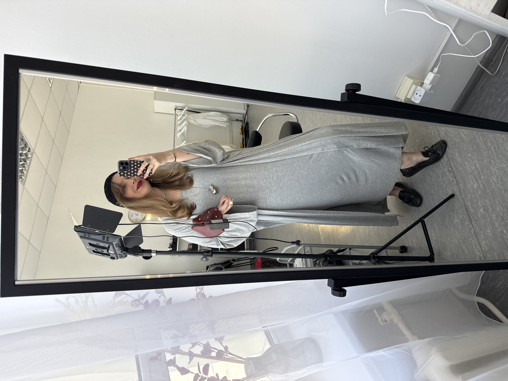 A woman in a gray dress and black loafers is taking a selfie in front of a mirror. She's wearing a black hat and holding a phone with a patterned case. The mirror has a black frame and stands on a black adjustable stand. A softbox light with a barn door attachment is visible behind her. In the background, there's a white wall with a visible electrical outlet and wiring, and a clothing rack with hangers and white garments. A white sheer curtain is draped near the bottom of the frame. The location is not specified as Izhevsk, Russia, and cannot be determined from the image alone.