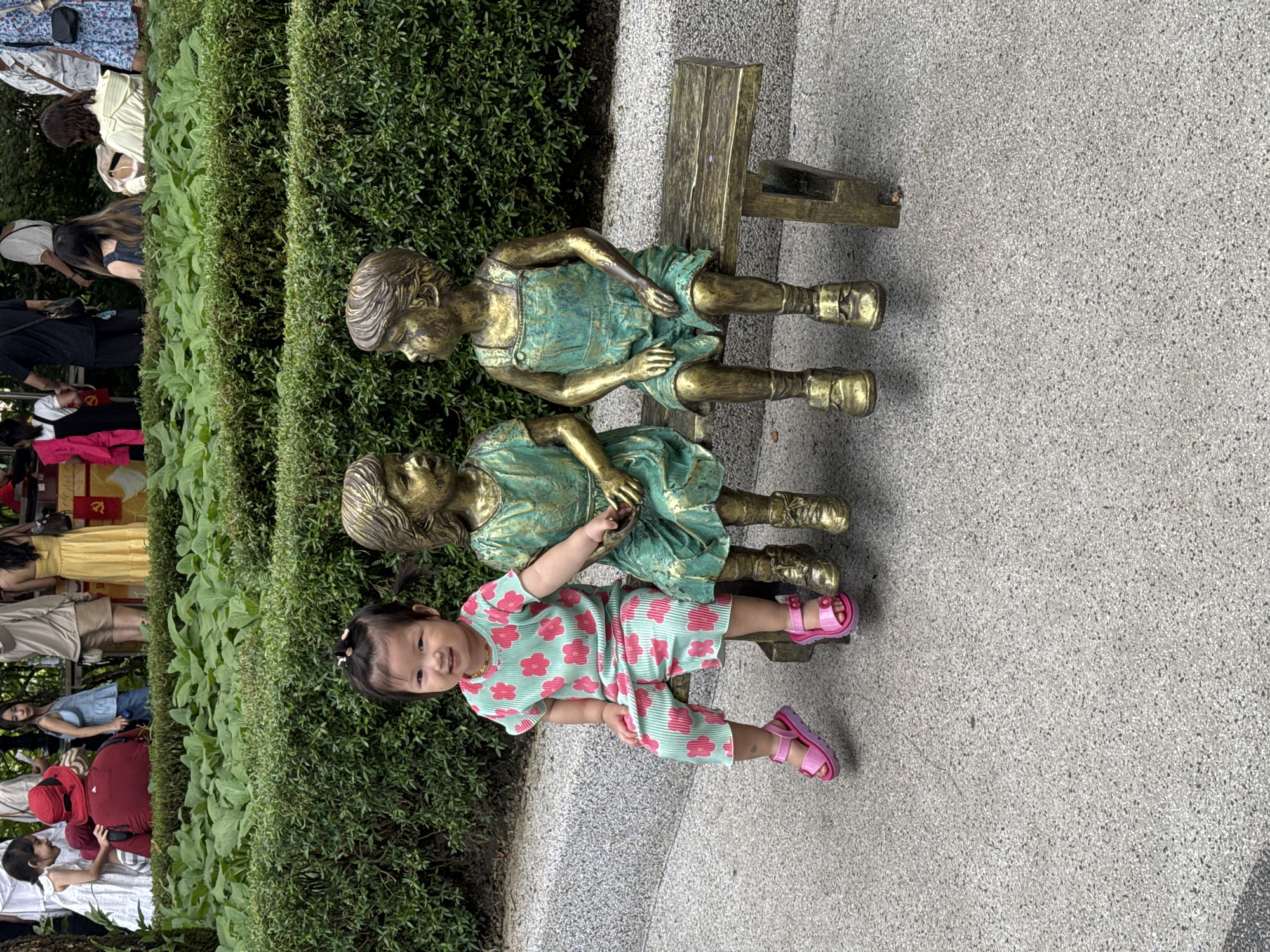 The image, oriented vertically, depicts a young child alongside two bronze statues in what appears to be a public park or garden in Đà Nẵng, Vietnam.
The central subject is a smiling toddler with dark hair, wearing a light green romper adorned with pink floral patterns and pink sandals. The child looks directly at the camera with a joyful expression. To the child's right are two bronze statues of young girls, depicted sitting on a wooden bench. The statues show signs of patination, giving them a green-tinged metallic appearance. They are dressed in what appear to be frocks or overalls.
Behind the child and the statues is a lush, well-maintained green hedge. Beyond the hedge, in the mid-ground, several individuals are visible, walking or standing, suggesting a public and active environment. These people are casually dressed, with various colors like white, yellow, blue, and red visible in their attire. The ground in the foreground is a light grey, speckled paved surface.
In the far background, a red banner with a yellow symbol, possibly a stylized hammer and sickle, can be faintly discerned, providing a subtle cultural cue relevant to Vietnam. The scene is brightly lit, indicating daytime, and the overall mood is cheerful, primarily due to the child's smile.