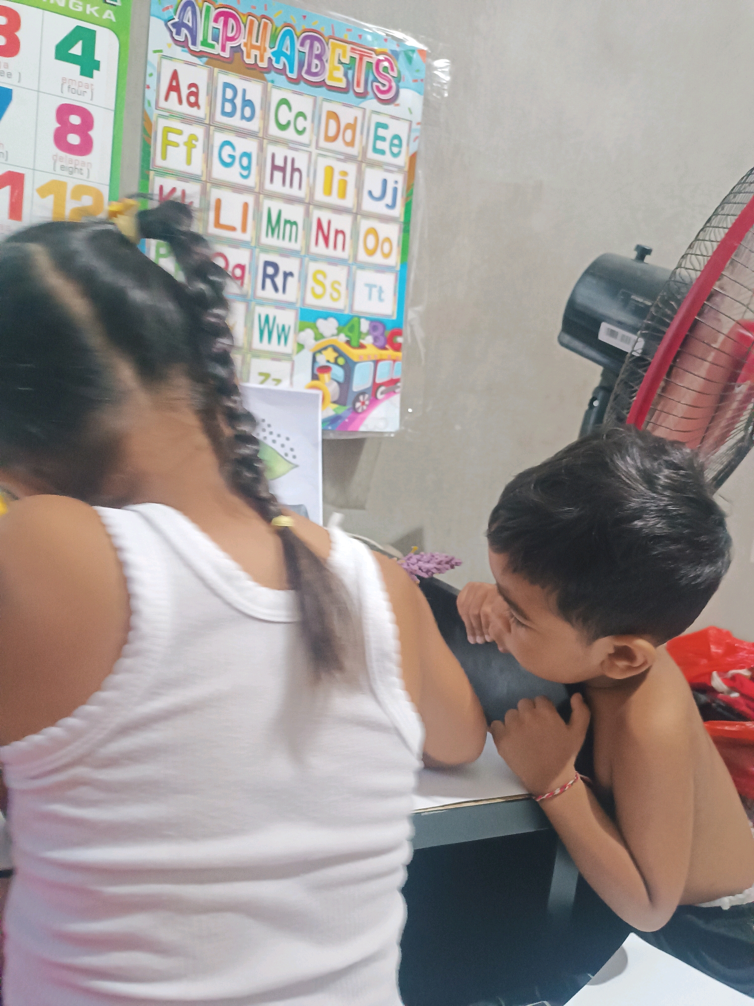 In a room, likely a home in Tabanan, Indonesia, two young children are engaged in an activity at a table. A girl, facing away from the camera, wears a white tank top and has her dark hair styled in braids. She is seated on the left. To her right, a boy, also seated, is shirtless and appears to be looking down at something on the table, possibly interacting with a toy train depicted on an alphabet chart. He is wearing a bracelet on his left wrist. The background features an alphabet chart with colorful letters and numbers, suggesting an educational setting. A black and red electric fan is visible to the right of the boy. The overall impression is one of a domestic scene where children are learning or playing.
