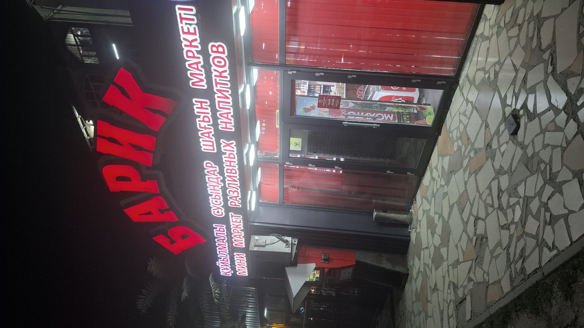 The image shows a storefront at night in Shymkent, Kazakhstan. The dominant feature is a large red neon sign that reads "БАРИК" in Cyrillic script. Below this, a smaller neon sign in a lighter color displays text in both Cyrillic and Latin alphabets, reading "ҚҰЙЫЛМАЛЫ СУСЫНДАР ШАҒЫН МАРКЕТІ" and "МИНИ МАРКЕТ РАЗЛИВНЫХ НАПИТКОВ". This translates to "Bottled Drinks Small Market" and "Mini Market for Draft Drinks" respectively.
The storefront itself has large glass windows and doors, with red vertical blinds covering portions of the windows. Inside, shelves with bottles and other products are visible. A refrigerated display case is positioned near the entrance. To the left of the entrance, an outdoor air conditioning unit is mounted on the building, and a red vending machine or service kiosk is situated on the pavement. The ground in front of the store is paved with irregular, interlocking stone tiles. The overall impression is that of a small convenience store or beverage shop operating in the evening.