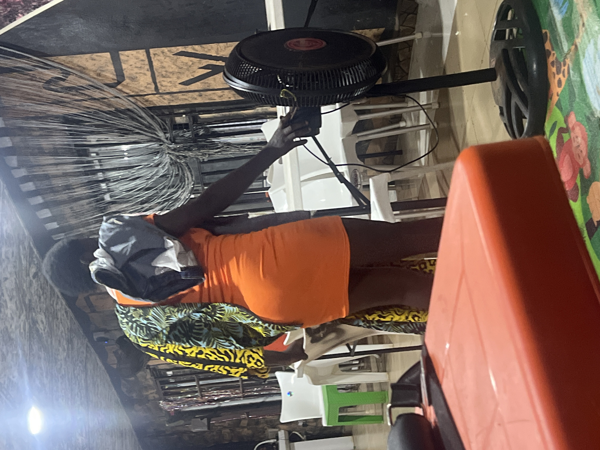 The image depicts an indoor or covered space in Obosi, Nigeria, likely in the evening or at night given the artificial lighting. A person, appearing to be female with dark skin, stands centrally in the frame. She wears an orange dress or top, a skirt with a vibrant yellow, black, and blue pattern, and carries a backpack over her left shoulder. Her right hand is positioned near the control panel of a large black standing fan, suggesting she might be adjusting it or holding something.
The scene appears to be a casual, functional environment, possibly a shop, workshop, or a simple seating area. In the immediate foreground, a large orange rectangular object, likely a table or counter, occupies the lower right portion of the image. To the right of the fan, a colorful mat or wall hanging featuring cartoon animals, including a giraffe and monkeys, is visible. Several white plastic chairs and a green stool are scattered in the background. On the far left, bundles of thin wires or cables are stacked, implying the business might deal with electrical supplies or similar materials. The ceiling is made of what appears to be unfinished plywood with dark beams. A bright light source is visible on the left side of the image. No discernible text is clearly visible. The overall mood is neutral, with no strong emotions conveyed.