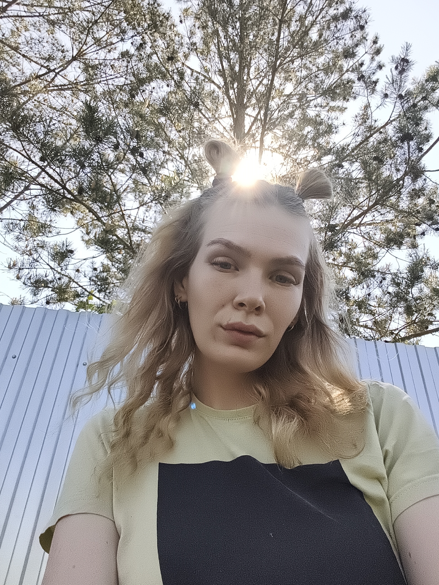 The photograph shows a person with long blonde hair styled in two small buns on top of the head, with the rest of the hair loose around the shoulders. The person is wearing a t-shirt that is light green in the upper part and has a dark blue block covering the chest area. Above the person, there is a tree with many branches reaching upwards, with the sun shining through the branches creating a bright spot in the center. In the background, there is a fence constructed of vertical corrugated metal panels. The location is Русский Вожой, Russia, as it has been provided.