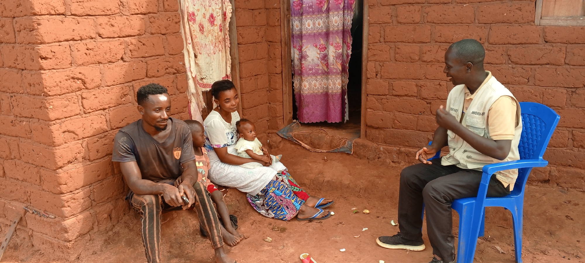 A scene in Ngalula, Democratic Republic of the Congo, depicts a family group near a brick building. A man with a beard sits on the ground next to a young child. A woman sits near them, holding a baby. Across from them, another man wearing a vest sits on a blue plastic chair. The background consists of brick walls and doorways with curtains. Debris is on the ground.