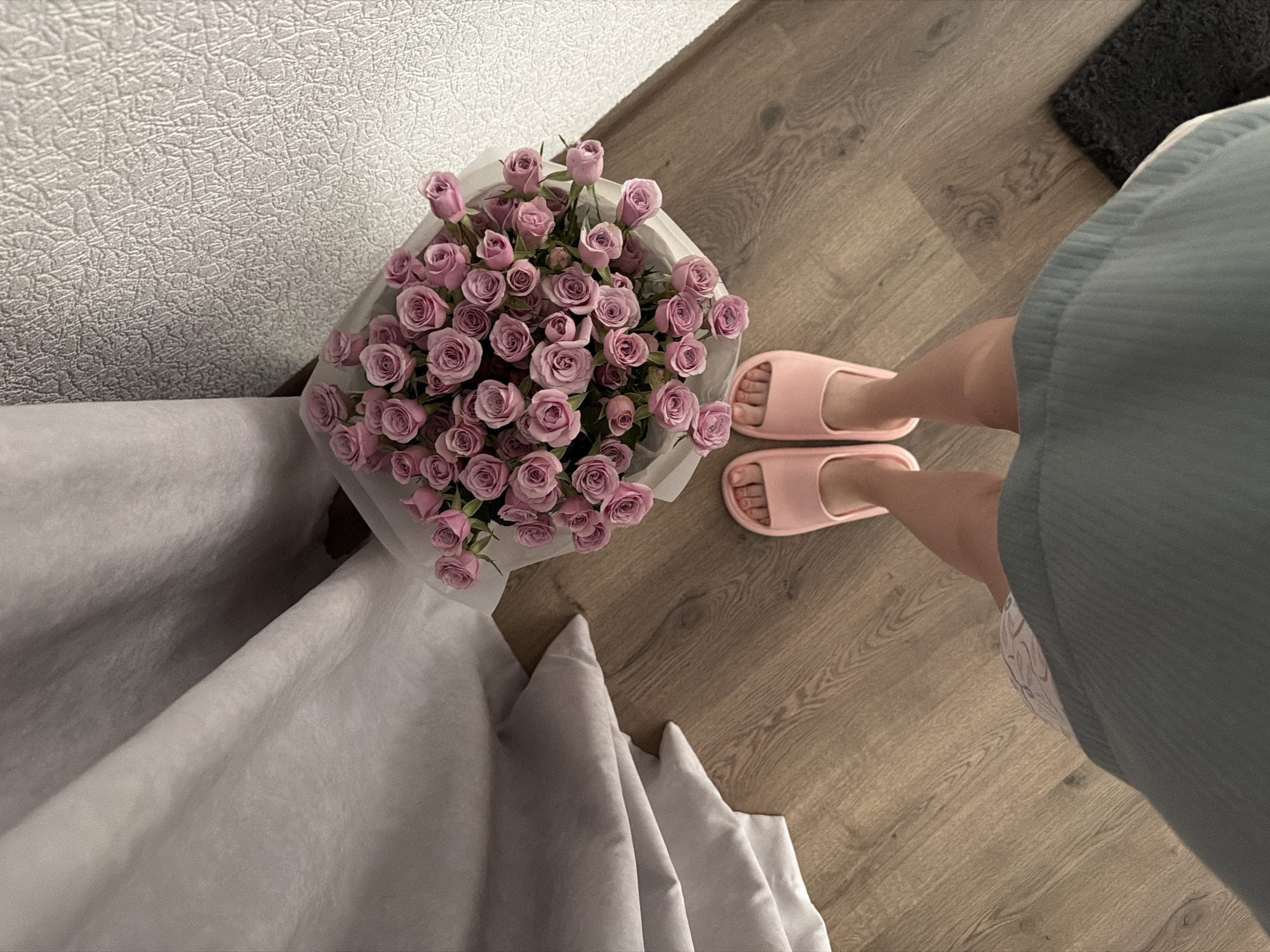 A first-person perspective shot shows a pair of feet wearing pink slippers, standing on a wooden floor. To the right of the feet is a bouquet of pink roses wrapped in white paper. Behind the bouquet is a textured white wall and to the left is a draped gray fabric. The overall scene suggests an indoor setting.