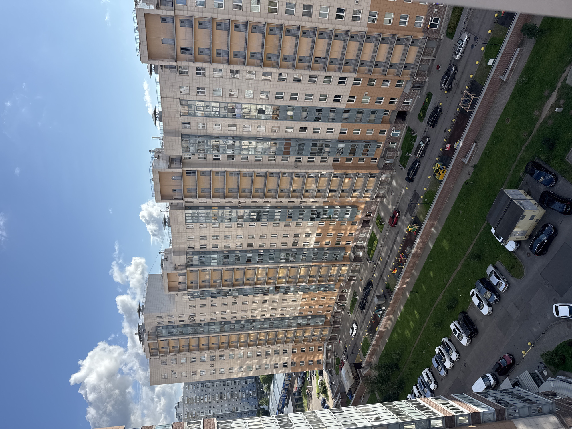 An aerial view captures a modern residential complex in Ladozhskoe Ozero, Russia, under a bright blue sky with scattered white clouds. The buildings are tall, multi-story structures with beige and light brown facades, featuring numerous windows and balconies. A street runs alongside the buildings, lined with parked cars of various colors, including white, black, silver, and red. Green grass and trees are visible in patches between the buildings and along the street. A playground with colorful equipment is situated near the street. The overall scene depicts a well-developed urban residential area.