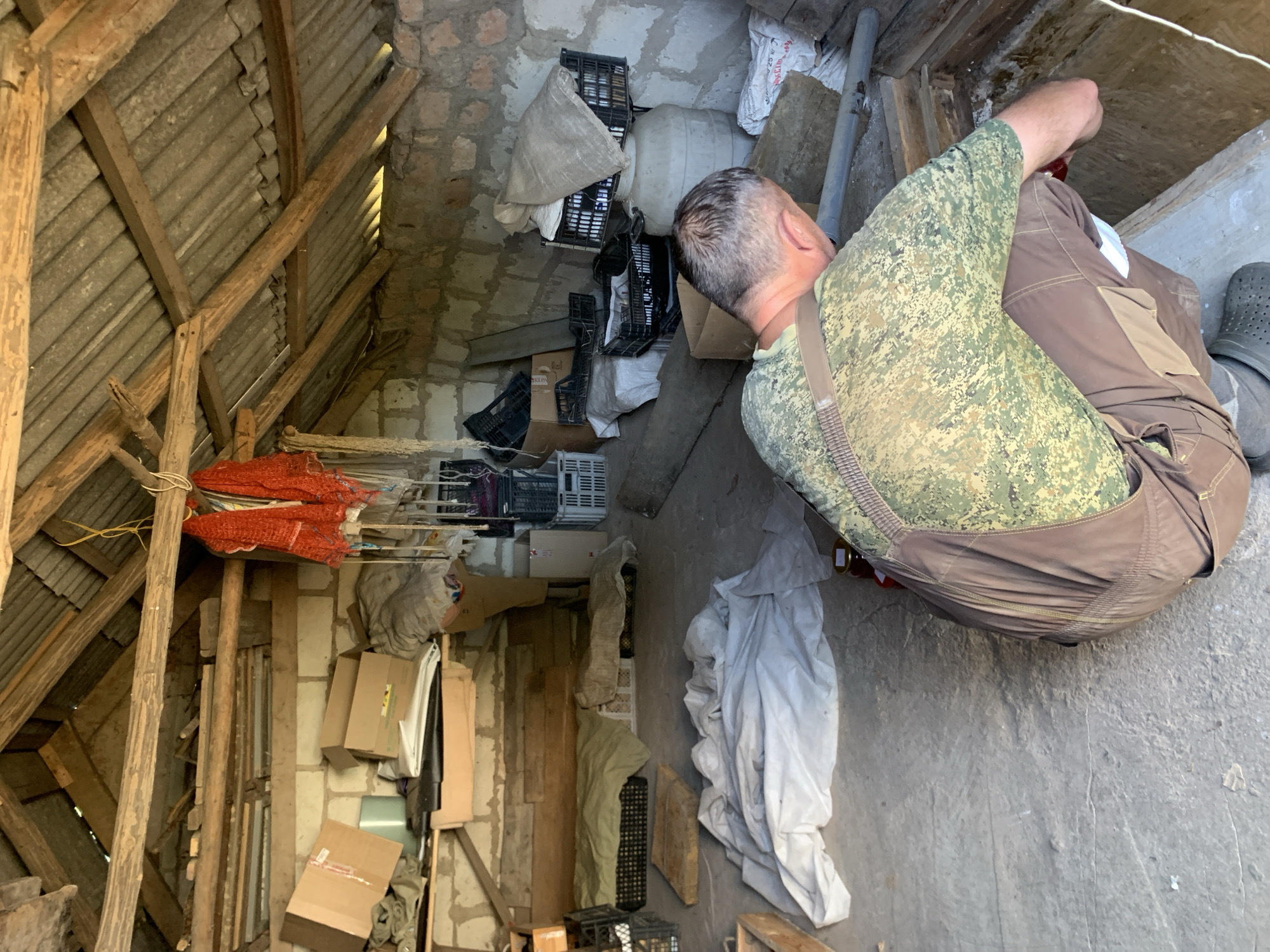 In a dilapidated shed with a corrugated metal roof supported by rough-hewn wooden beams, a man in camouflage overalls and a patterned shirt crouches. He is positioned in the foreground, facing away from the viewer and towards a wooden structure at the right rear of the shed. The shed is cluttered with various items, including cardboard boxes, plastic crates, sacks, and large white plastic containers. A white sheet or tarp is draped on the floor in the left-center of the image. The walls appear to be constructed from concrete blocks. The scene is dimly lit, suggesting it might be daytime with indirect light entering the shed. The man's focus is directed downwards as he appears to be working on or examining the wooden structure. The overall impression is one of disarray and storage within a rustic, possibly agricultural, setting.