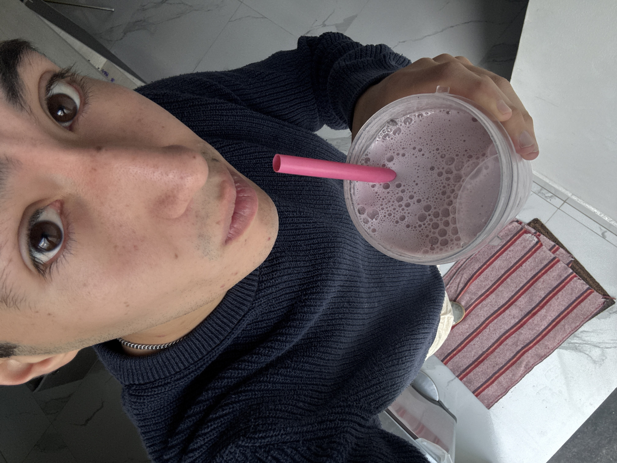 A close-up, slightly tilted, high-angle shot depicts a person holding a clear plastic cup filled with a pinkish-purple beverage, likely a smoothie or shake, containing numerous bubbles. A bright pink straw is inserted into the drink. The person is wearing a dark blue knitted sweater and a thin silver chain necklace. Their face is partially visible, showcasing brown eyes and light skin with some visible pores and stubble. The background includes a marble-like surface, possibly a countertop or floor, and a striped red and beige dishtowel. The location is not discernible from the image alone. San Bernardino Tlaxcalancingo, Mexico is not identifiable within the image.