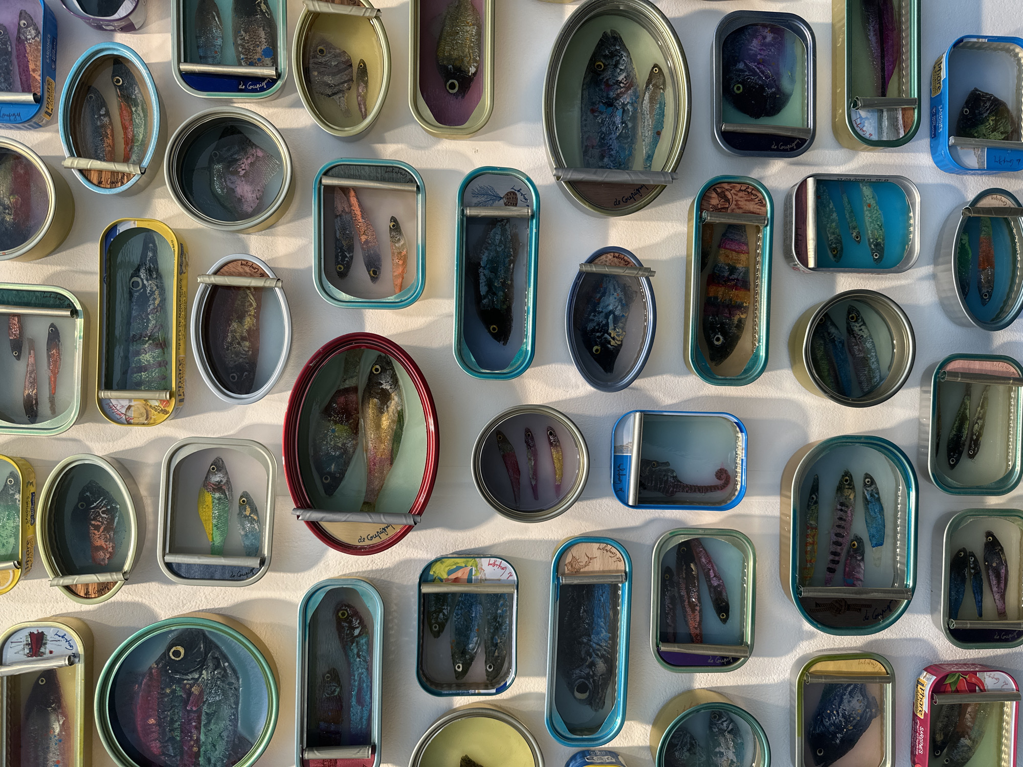 The image shows a wall adorned with numerous sardine tins. Each tin has been repurposed into a miniature art display, housing a painted or sculpted depiction of fish inside. The sardine tins vary in shape and size, with both circular and rectangular options visible. The artwork within the tins also exhibits a range of styles and colors. Some tins have labels visible around their edges. There is nothing in the image that indicates it is in London, United Kingdom.