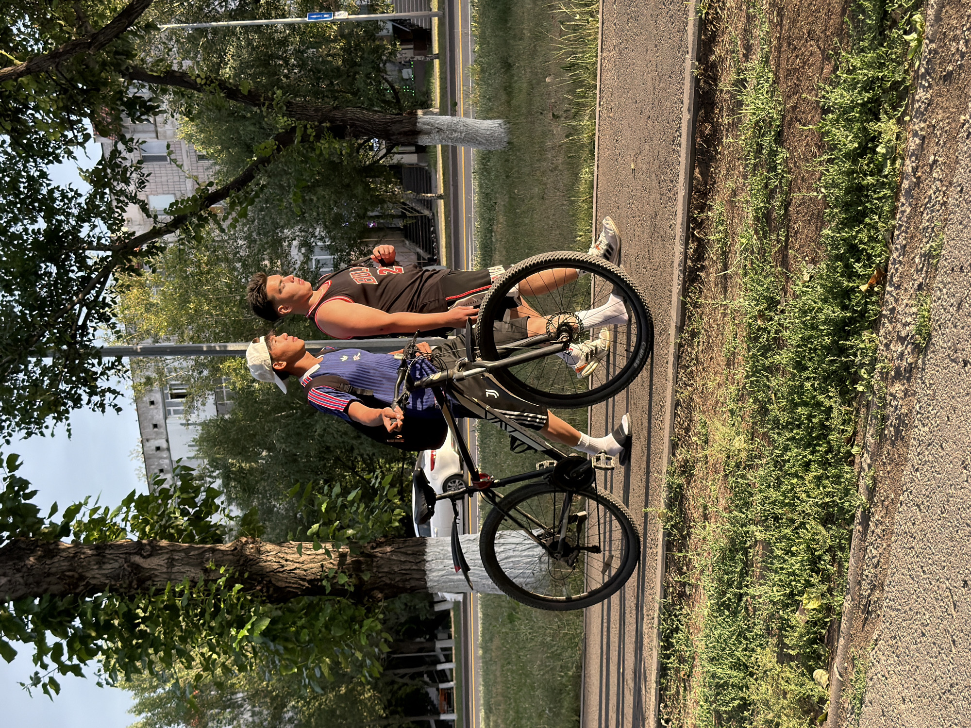 The image depicts two young men with a bicycle on a paved path next to a grassy area. The man in the foreground is wearing a white cap, a blue and white striped shirt, and dark shorts. He is looking up and to the right. The man behind him is wearing a black tank top with red trim and dark shorts. He is also looking up and to the right. Both men are wearing athletic shoes. The bicycle is black and appears to be a mountain bike. The scene is outdoors with trees and buildings in the background. The lighting suggests it is daytime, possibly late afternoon, with the sun casting shadows. The overall mood appears relaxed. The location is specified as Astana, Kazakhstan. There is a blue road sign visible in the upper left corner of the image. The building in the background appears to be an apartment complex.