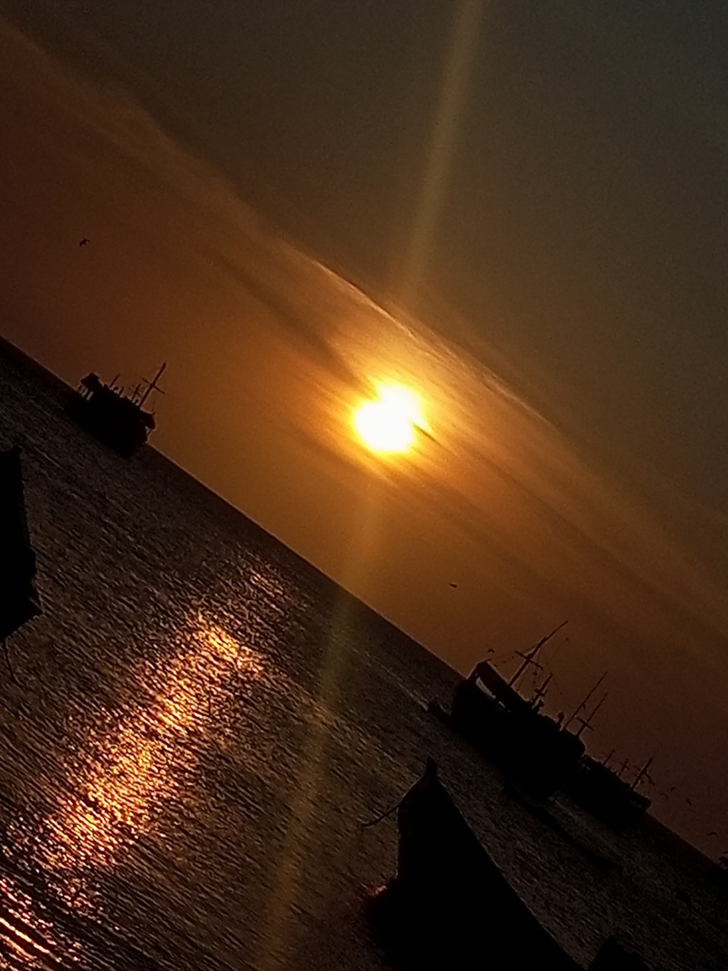 The image shows a seascape at sunset in Juan Griego, Venezuela. The sun is positioned near the horizon, its light reflecting off the water's surface. Several silhouetted boats are visible on the water.