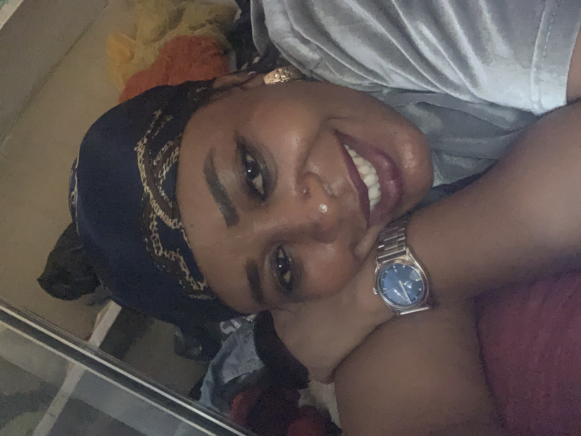A woman with dark skin and a dark head covering with a gold-colored pattern smiles at the camera. She has a silver watch on her left wrist. She appears to be lying down. The setting is undiscernible. The image does not provide any information to determine if the location is Sabon Tasha, Nigeria.