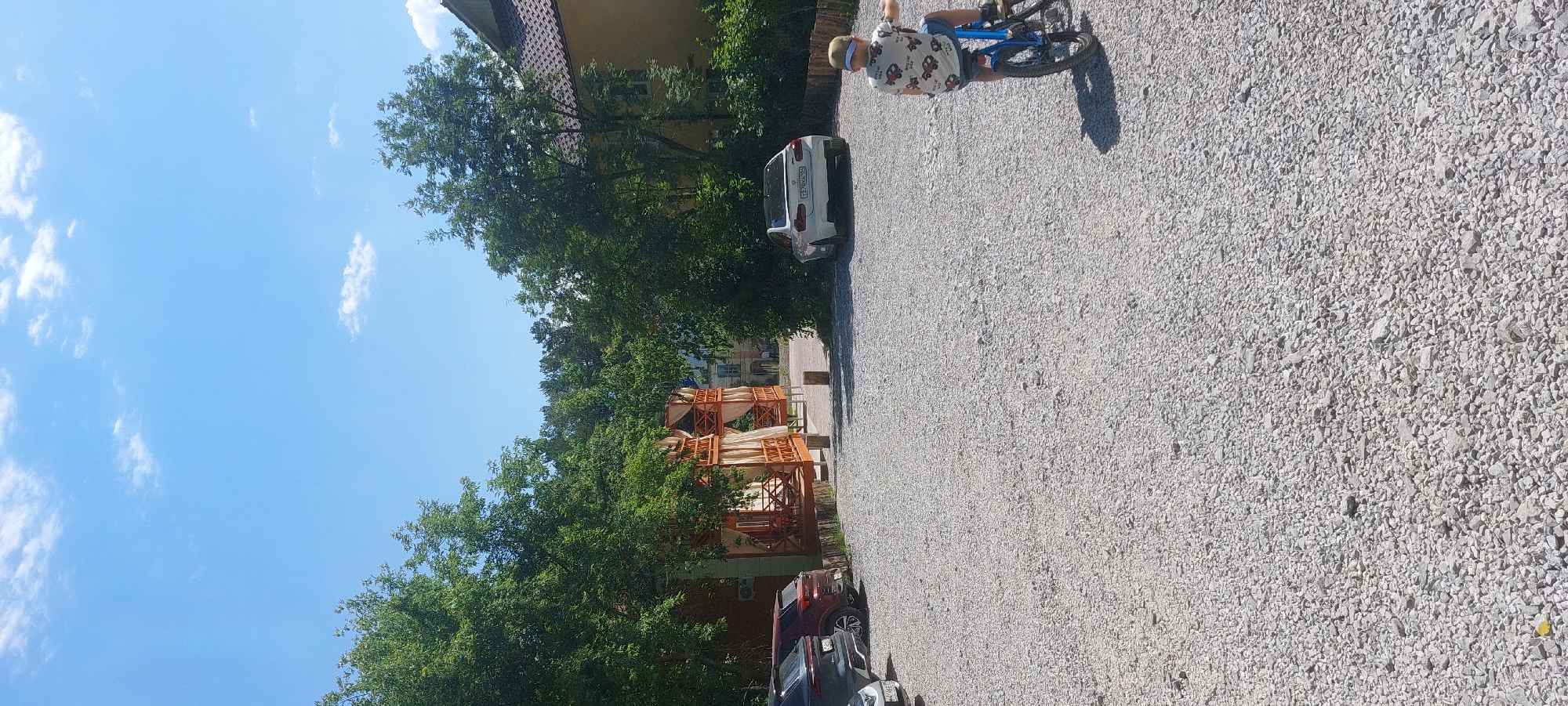 The image depicts a person riding a bicycle on a gravel surface, likely a parking lot or road, in Berdsk, Russia. The person, seen from the rear and side, is wearing a white t-shirt with a colorful pattern and blue shorts, and a light-colored cap. They are on a blue bicycle and are angled towards the right side of the frame.
In the background, a white car is parked. Further back, there are trees with lush green foliage and a building with a tiled roof. Two wooden structures with draped fabric are also visible, possibly gazebos or covered seating areas. The sky is a clear blue with a few white clouds. The scene appears to be set during daylight hours, likely a sunny day, given the shadows and bright lighting.
There is some visible text on the license plate of the white car, which reads "E570XA 194". The overall setting suggests an outdoor recreational area or a residential neighborhood. The activity is simple leisure; someone is enjoying a bicycle ride.