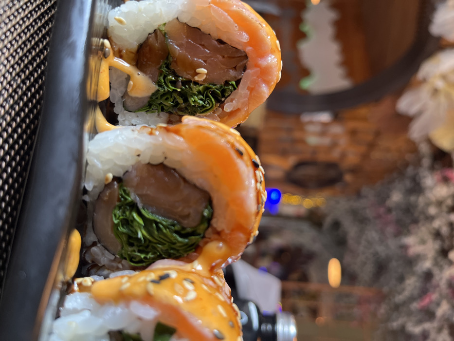 A close-up photograph depicts three sushi rolls arranged on a black rectangular plate, resting on a dark, woven placemat within a restaurant environment in Bad Sobernheim, Germany. Each sushi roll consists of white rice, topped with a slice of orange-pink salmon, glazed with a light orange-brown sauce, and sprinkled with white sesame seeds. The visible interior filling includes dark green leafy greens and a reddish-brown component, likely fish. A creamy, pale orange sauce is drizzled over the rolls and pools on the plate. The background is out of focus, revealing blurred elements of a restaurant interior, including wooden-like architectural arches, various bokeh lights (a distinct blue light and several yellow lights), white floral decor, dark foliage, and a partial view of a dark bottle with a silver cap. No individuals are present in the image.