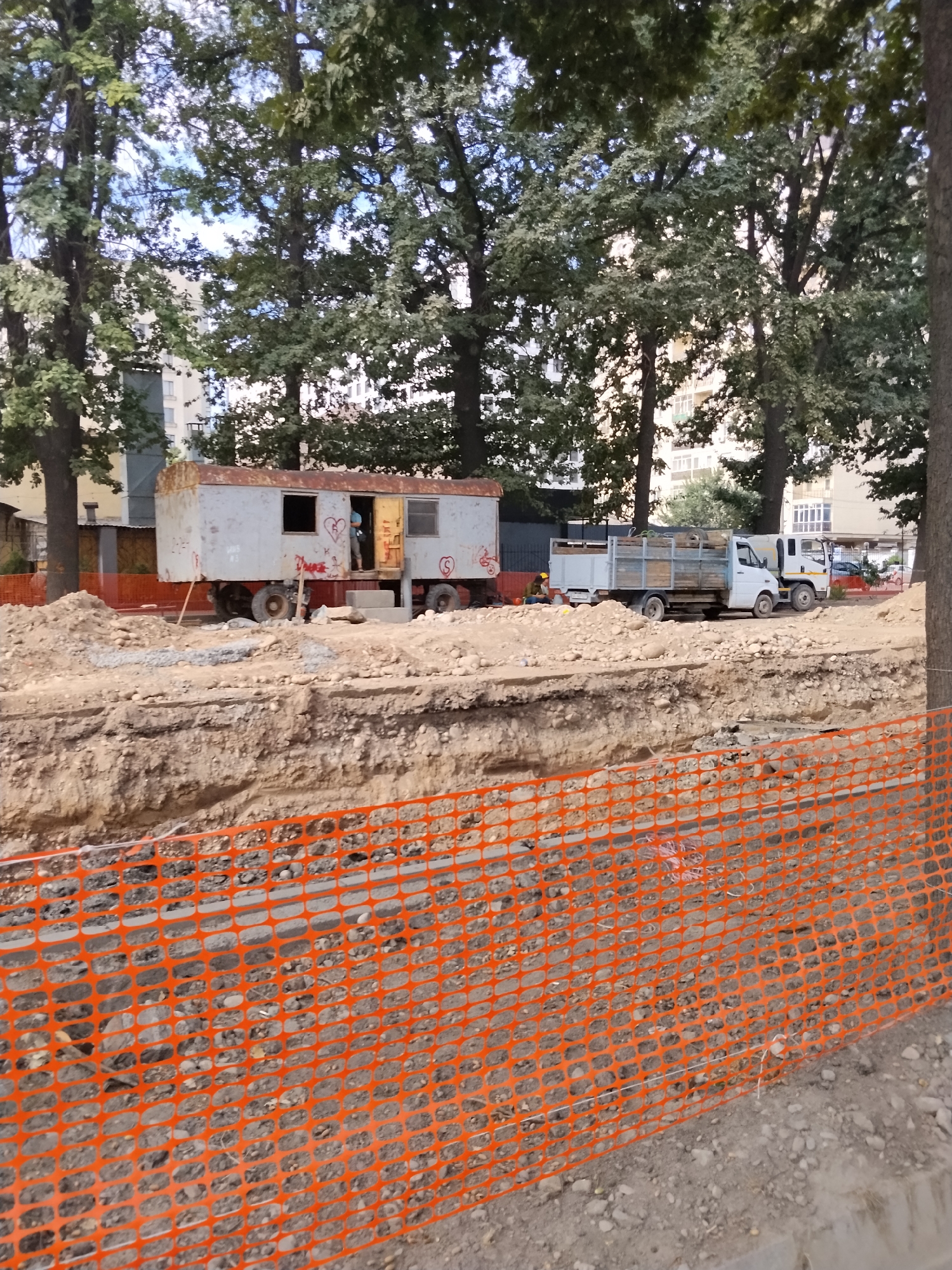 A construction site in Bishkek, Kyrgyzstan is depicted. In the foreground, an orange plastic safety net divides the frame. Behind the net, an excavated trench reveals layers of soil. The middle ground features a makeshift trailer with wheels, a person standing in the doorway, and graffiti on its exterior. Two trucks, one loaded with wooden planks, are parked on the uneven ground. In the background, several trees with lush green foliage tower over apartment buildings. The sky is visible through the leaves of the trees. The scene suggests ongoing infrastructure work.