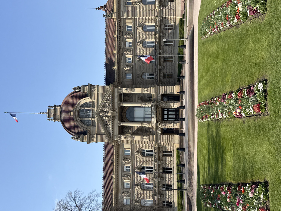 The image depicts the Palais du Rhin (Rhine Palace) located in Place de la République, Strasbourg, France, under a clear blue sky. The grand, symmetrical building is constructed from light-colored stone, featuring a central reddish-brown dome topped with a finial from which a French tricolor flag flies. The dome is adorned with statuary and intricate architectural details. Below the dome, the main facade includes a prominent portico with large classical columns and an arched central window, flanked by numerous rectangular windows across multiple stories. Another smaller French tricolor flag is mounted on a pole to the left side of the building's facade at a lower level. The building is topped with reddish-brown tiled roofs on its lateral wings.
In the foreground, a manicured green lawn is visible, featuring three distinct, rectangular flower beds arranged parallel to each other. These beds are filled with an assortment of vibrant flowers including shades of red, pink, white, purple, and yellow. A wide, multi-tiered set of stone steps leads up to the building's main entrance. A series of black bollards are evenly spaced along the base of these steps. Bare tree branches are visible on the far left edge of the frame. The scene is devoid of people or overt actions.