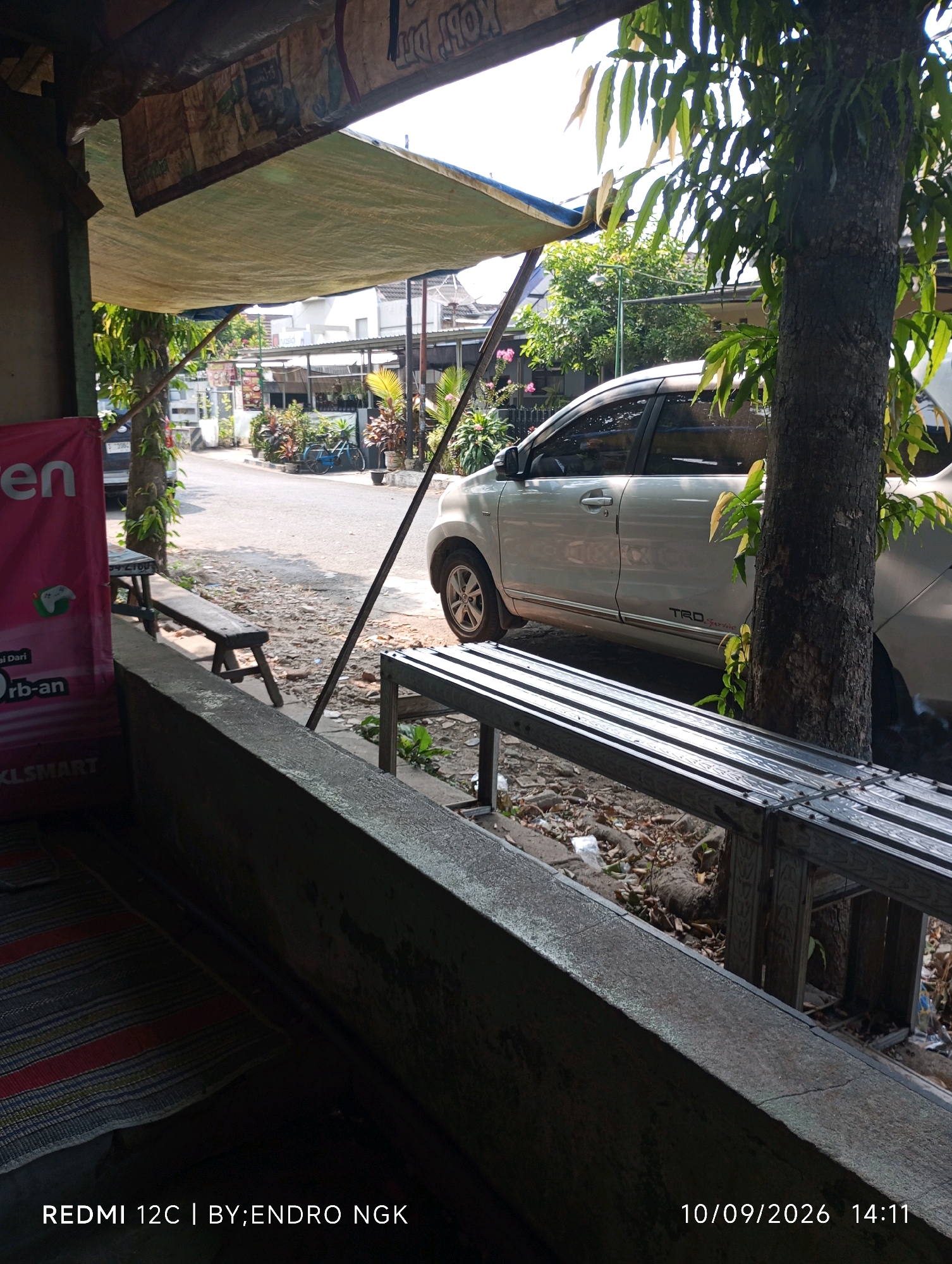 The image depicts an outdoor scene, possibly a roadside stall or a simple establishment, in Nganjuk, Indonesia. In the foreground, a textured concrete ledge dominates the lower portion of the frame, with a striped mat visible underneath it. To the left, a partially obscured pink banner displays text in what appears to be Indonesian, with a small icon resembling a video game controller. Behind this, a wooden bench rests on the ground, its surface weathered.
The midground reveals a dirt and gravel area with a few scattered leaves and debris. A silver car is parked on the right side of a paved road. The car has a sticker on its lower body that reads "TRD Sportivo". Next to the car, a large tree trunk stands tall, with its branches and leaves extending upwards and partially obscuring the sky. A metal bench, constructed from parallel metal bars, is situated in front of the tree.
Further in the background, across the road, are several buildings with awnings and a variety of plants and flowers, suggesting a commercial or residential area. A blue bicycle is visible leaning against something among the plants. The overall impression is a humble, sun-drenched street scene. The timestamp in the lower right corner indicates the image was taken on September 10, 2026, at 2:11 PM. The text at the bottom left identifies the device as "REDMI 12C" and the photographer as "BY:ENDRO NGK".