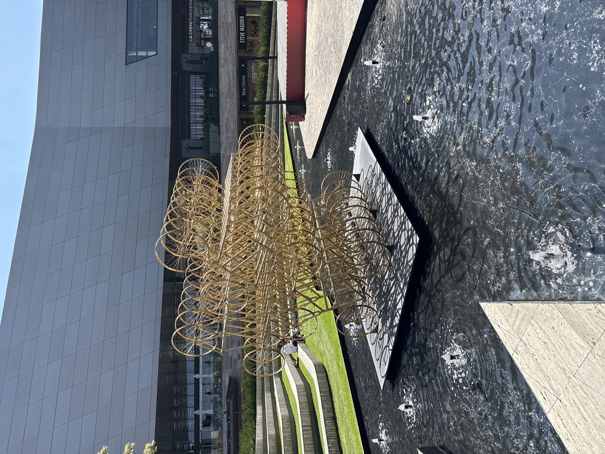 The image captures an outdoor architectural scene in Mexico City, Mexico, featuring a large, abstract, golden-colored sculpture composed of interconnected circular shapes. The sculpture stands next to a water feature, the dark, rippled surface reflecting light and containing small fountains. Adjoining the water feature is a strip of manicured green grass bordered by concrete structures. In the background, there's a modern building with a grey facade and large glass windows. Adjacent to the building are storefronts featuring names like "MacStore" and "Steve Madden," with the sign of "La Crepe Parisienne." A person stands on the grass area beside the sculpture.