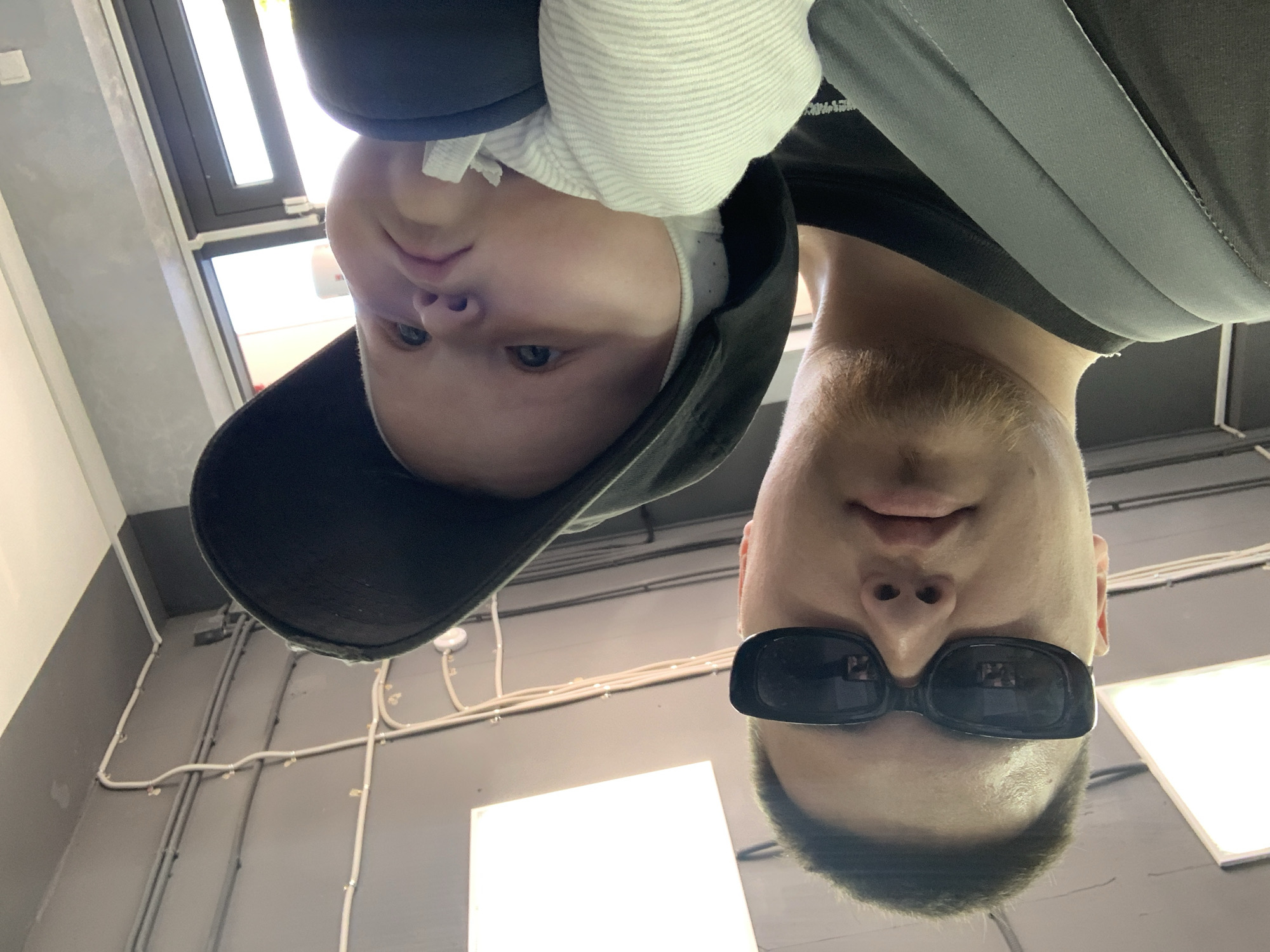 A man wearing black sunglasses and a black baseball cap, possibly in Stavropol, Russia, holds a baby in a carrier. The baby is wearing a white, striped onesie and a white bonnet. The background shows a ceiling with fluorescent lights and exposed electrical wiring, suggesting an indoor public space such as a shopping mall or community center.