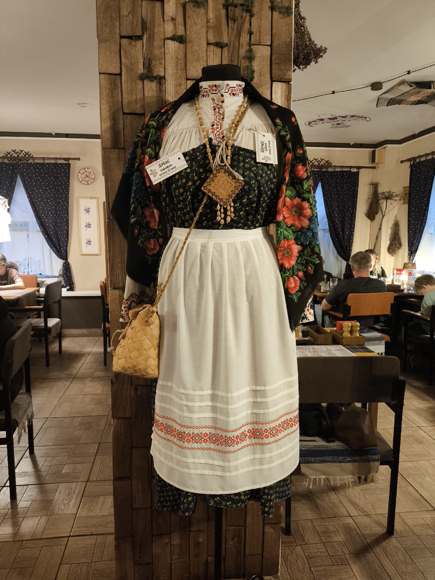 The image depicts a mannequin dressed in traditional Russian clothing, displayed in what appears to be a restaurant or cultural establishment in Perm, Russia. The mannequin is adorned with a dark floral dress, a white apron with embroidered trim, a colorful floral shawl, and a beaded necklace with a wooden pendant. A woven straw bag hangs from its shoulder. The background shows a rustic interior with wooden beams, patterned curtains, and diners seated at tables. The lighting is warm, suggesting an indoor setting. The overall impression is one of cultural preservation and display within a public space.