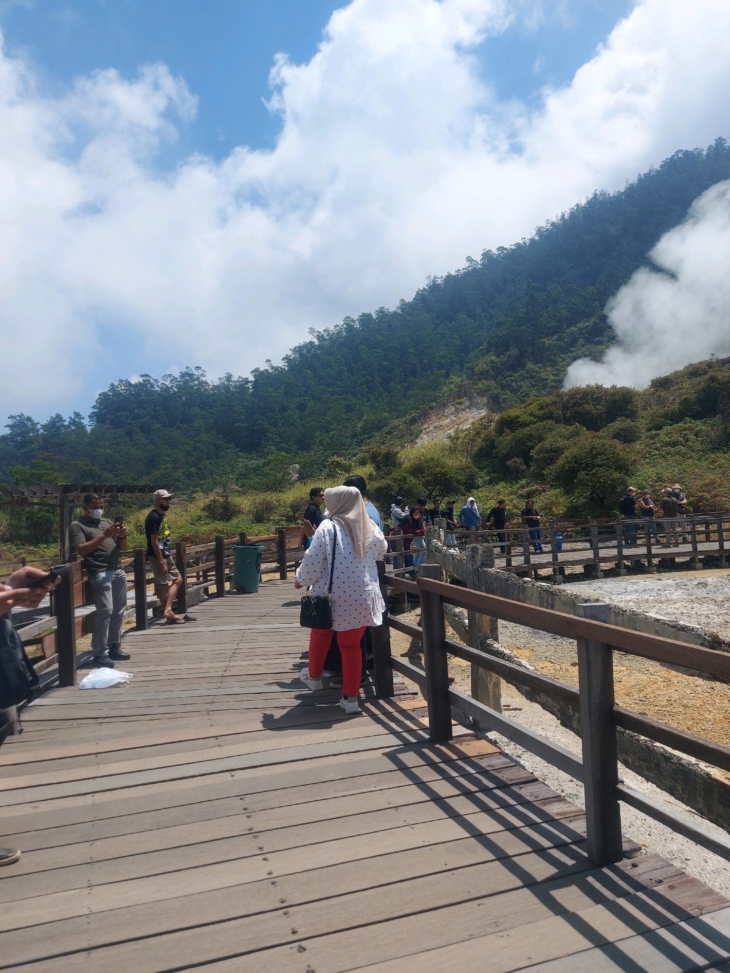 The image depicts a scene at the Sikunang crater in Indonesia, a location characterized by its volcanic activity. A wooden boardwalk has been constructed to allow visitors to safely observe the crater. Several people are walking on the boardwalk, some are standing and looking at the crater, and others are taking pictures. The environment shows a mountainous landscape covered in lush green vegetation, with a visible plume of steam rising from the crater. The sky is partly cloudy with a clear blue sky visible in some areas. The overall impression is that of a tourist attraction where visitors are experiencing a natural geological wonder.