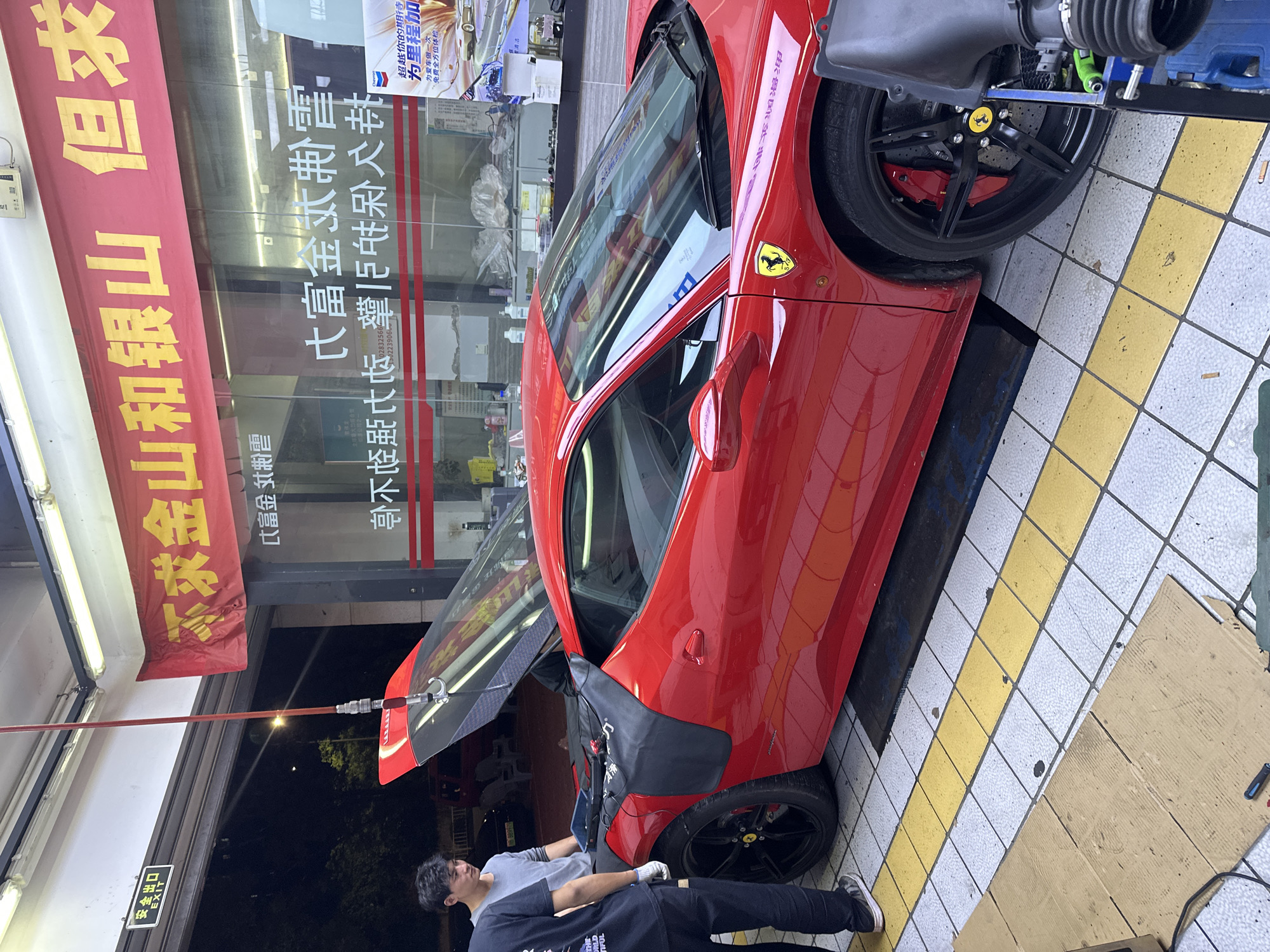 The image captures an evening or nighttime scene outside a car service center in Chongqing High-tech Zone, China, where a bright red Ferrari sports car is undergoing maintenance. The car, possibly a Ferrari 458, is parked on a tiled surface with white and yellow squares, partially extending into what appears to be a garage bay. Its front hood or engine bay is open, revealing internal components and connected equipment, including a black ribbed hose that might be part of an air intake system. A long red support pole extends from above to the open panel, and a protective black covering is draped over the car's body near the front wheel.
Two men, likely technicians, are working on the vehicle. One man in a grey t-shirt stands near the car's front, looking into the engine bay. Another man in a dark t-shirt and pants stands closer to the foreground, observing the work. The Ferrari's black wheels with red brake calipers and the distinctive prancing horse emblem on the wheel hubs and side panel are clearly visible.
The background features a building with large glass doors and windows, reflecting lights and showing various signs inside. A prominent red banner with yellow Chinese characters hangs vertically on the building. Visible text on the banner and through the glass includes slogans such as "不求金山和银山 但求..." (Not seeking gold mountains and silver mountains, but seeking...), "富金 制震" (Fù jīn zhì zhèn - related to vibration damping), "超越你的期待" (Beyond your expectations), "为里程加" (Add to mileage), and "为爱车做一次 免费全方位体检" (Give your beloved car a free comprehensive check-up). A partial phone number "1302239060" is also visible. An "安全出口" (Safety Exit) sign with "EXIT" in English is mounted above a doorway. The artificial lighting suggests the late hours of the day.