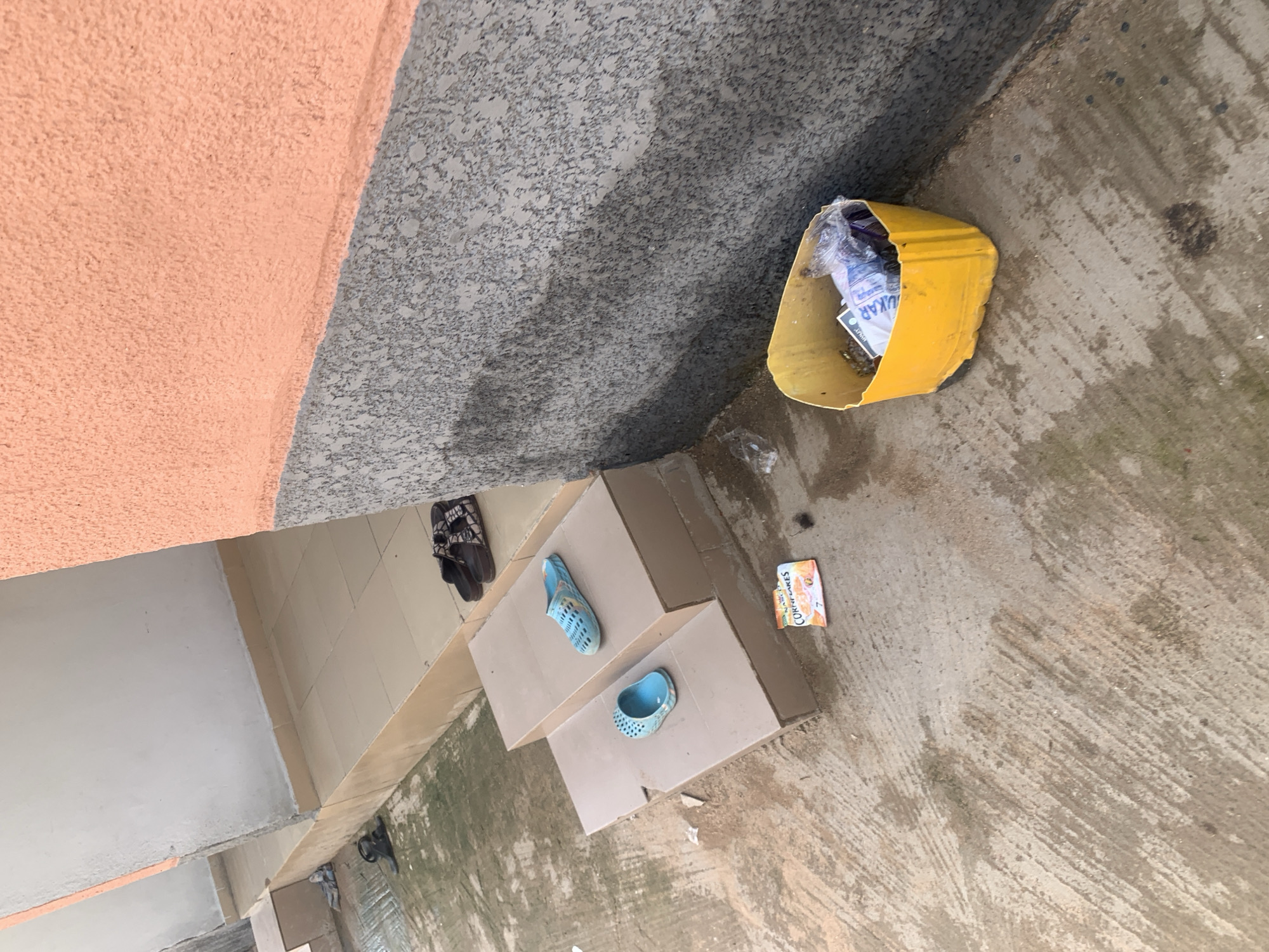 The image depicts an outdoor scene on what appears to be a pathway or concrete area, possibly in front of a building. The ground is wet and shows some wear and tear, with visible water stains and debris.
In the foreground, a bright yellow trash can is overflowing with assorted items, including plastic bags and what appear to be papers. To the left of the trash can, on a set of tiled steps, are two pairs of blue slip-on sandals. Further up the steps, another pair of dark-colored sandals is visible.
A small, brightly colored packet, possibly food packaging, lies on the ground near the steps. The background reveals a building with an orange and gray textured wall, and a light-colored tiled walkway leading to doorways. The scene suggests a somewhat unkempt outdoor area, possibly a common space associated with residential units or a public building.
Based on the provided information, this scene is located in Jauro Yinu, Nigeria.