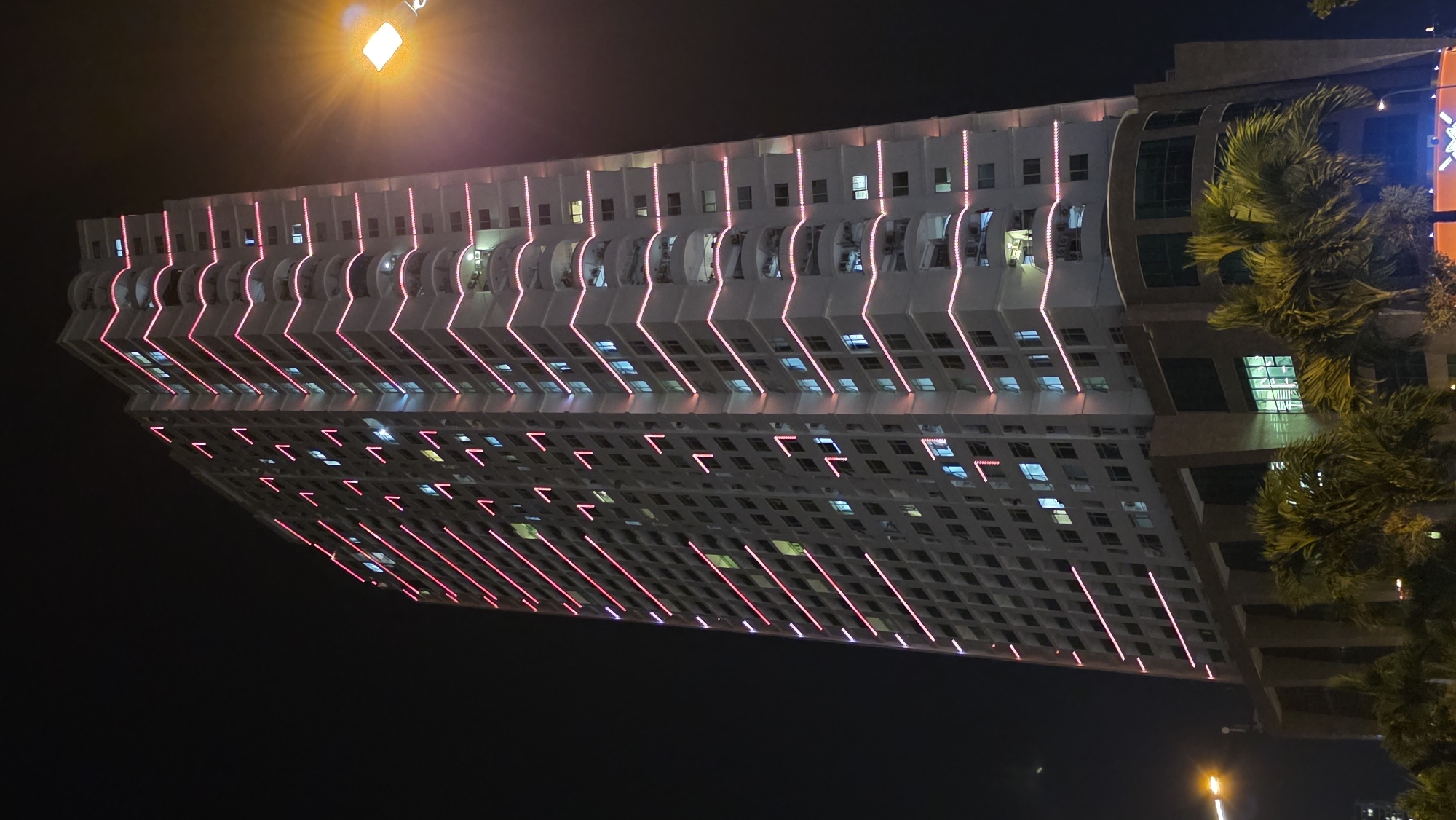 The image depicts a high-rise building at night, presumably in Nha Trang, Vietnam. The building is brightly lit with pink neon lights outlining its architectural features. Windows are visible across the facade, with some illuminated by interior lights. Palm trees are present on the right side of the frame, suggesting a coastal location. Street lights are visible in the background. The overall environment is urban and illuminated at night.