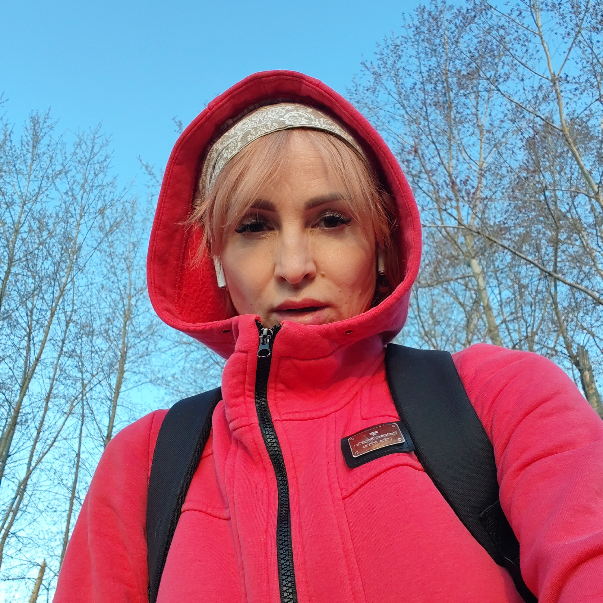 A woman is depicted in a selfie, wearing a red hooded sweatshirt with a black backpack. She has light pink hair, a patterned headband, and white wireless earbuds. The zipper of her sweatshirt is partially zipped. The background includes a clear blue sky and bare trees. The picture appears to be taken outdoors. The location is assumed to be in the city of Chaikovsky, Russia.