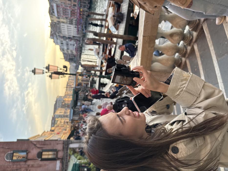 An outdoor, wide-angle image depicts a bustling canal scene in Venice, Italy, at golden hour. In the immediate foreground, a young adult female with long brown hair, wearing a beige trench coat, is positioned behind a light-colored stone balustrade. She holds a black smartphone in her left hand and a black digital camera in her right, aiming the camera towards the background with a focused, slightly strained expression.
The midground and background reveal a wide canal lined with historic, multi-story buildings featuring varied facades and traditional windows. A crowded pedestrian walkway runs along the canal, populated by numerous individuals walking or seated at outdoor cafe tables beneath green awnings. A prominent black street lamp with three lanterns stands on the right side of the walkway. Several boats are moored to numerous wooden poles within the canal. The sky above displays soft orange and blue hues. A partial view of another person in light blue jeans and a brown jacket is visible to the right of the main subject.