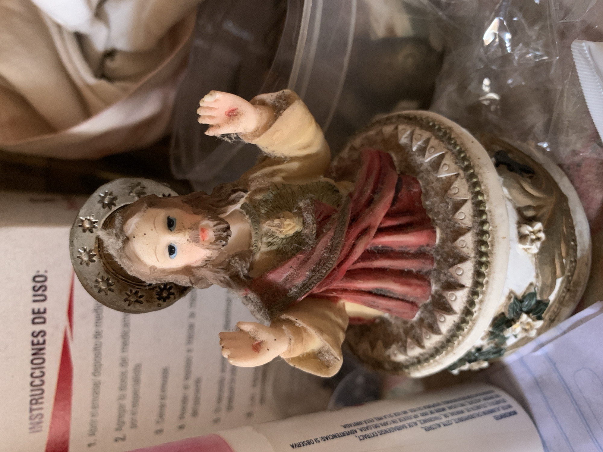 The image displays a religious figurine depicting the Sacred Heart of Jesus, situated in an indoor setting in Santa Ana, Venezuela. The figurine features Jesus with blue eyes, a brown beard, and long brown hair, wearing a beige tunic and a red cloak. A halo with star-like patterns encircles his head, and the Sacred Heart is visible on his chest. His outstretched hands show a faint red mark on the right palm, indicating a stigmata-like representation. The figurine rests on an ornate round base decorated with geometric patterns and small floral elements in green and white.
A notable detail is the accumulation of dust or lint on the figurine's surface, particularly on the clothing and hair, suggesting it may have been stored or not recently cleaned. The image itself is rotated, making the figurine appear horizontally oriented.
The background, out of focus, reveals various household items, including light-colored fabric, clear plastic packaging, and multiple papers. Visible text on one of the papers is in Spanish and includes "INSTRUCCIONES DE USO:" (Instructions for use), followed by steps like "1. Abrir el envase deposito de medic" (1. Open the container/deposit of medicine) and "2. Agregar la dosis del medicam" (2. Add the dose of medicine). Other fragments read "ALOE BARBADENSIS EXTRACT," and "ADVERTENCIAS: SI OBSERVA" (WARNINGS: IF YOU OBSERVE), alongside "APREL ESTA IRRITADA" (APREL IS IRRITATED), suggesting product instructions or warnings. A pink-capped object, possibly a tube or bottle, is also partially visible. The scene is a still life, set indoors with soft, possibly natural, lighting.
