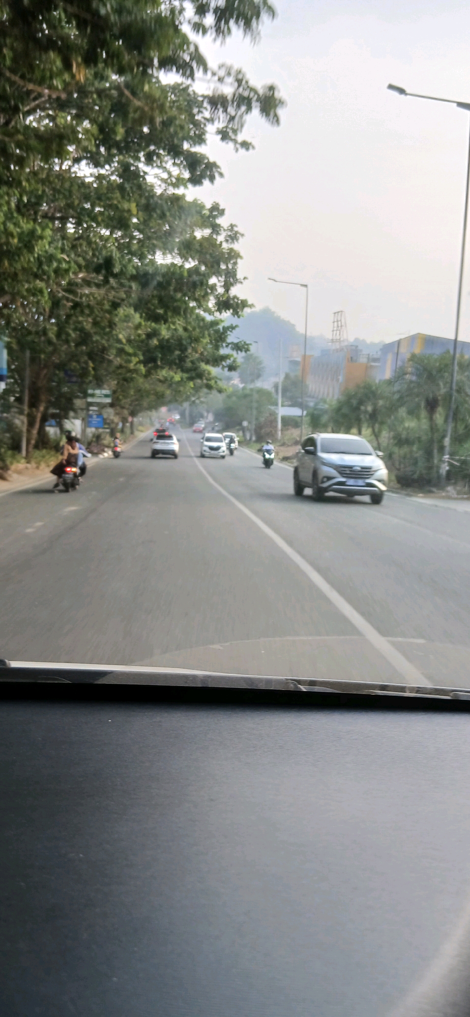 The image captures a street scene in Samarinda, Indonesia, likely during daytime. The viewpoint is from inside a vehicle, looking through the windshield, which is partially visible at the bottom of the frame. The road appears to be a multi-lane highway or main road, with white lane markings.
On the road, several vehicles are present. In the foreground, a silver SUV is driving in the right lane. Further down the road, there are at least two white cars and a motorcycle. On the left side of the road, a motorcycle with two occupants is also visible, driving in the same direction as the other vehicles. The road is flanked by trees on the left and right, with lush green foliage.
In the background, hills or mountains can be seen, partially obscured by a hazy sky. There are also structures in the distance, including what appears to be industrial buildings or a factory complex with yellow and blue elements, suggesting an urban or semi-urban industrial area. Streetlights are visible along the roadside. The overall atmosphere suggests a typical day of traffic and transportation in the city.
